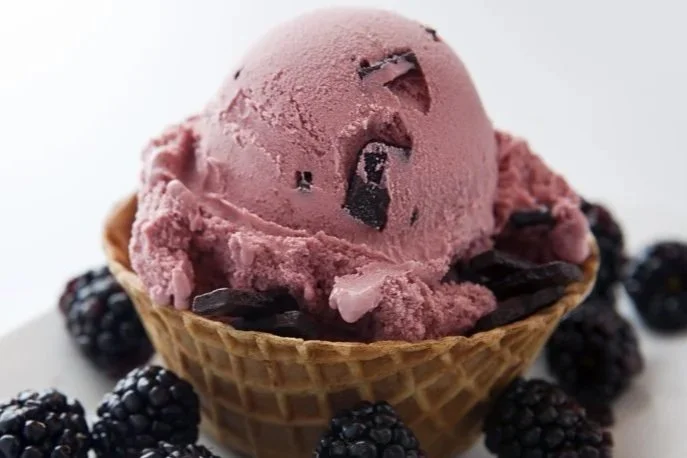 Black Raspberry Chocolate Chip ice cream scoop with chocolate chunks in a waffle cone, surrounded by black raspberries.