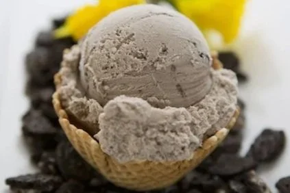 A scoop of grayish ice cream in a waffle bowl, topped with a gray cookie and garnished with yellow flowers and chocolate cookies in the background.