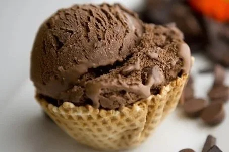 Close-up of a chocolate ice cream scoop in a waffle cone with chocolate pieces in the background.