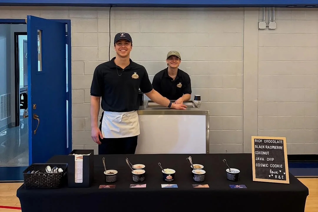 Two smiling people, a man and a woman, stand behind a table with different flavors of ice cream in small bowls. The man wears a black shirt, baseball cap, and apron, and the woman wears a black shirt and cap. A sign on the table lists ice cream flavo