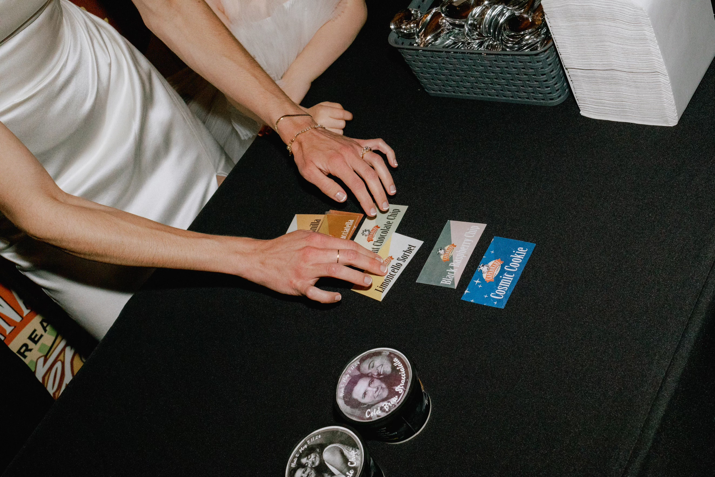 Two women with long nails and jewelry sit at a table sorting through colorful vintage-style card tickets and two small cups with black and white photos of a couple, labeled 'Cold & Stormy' and 'Matrimonial' at a wine or liquor tasting event.