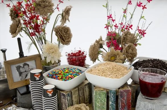 A dessert table with bowls of candy, crushed cookies, and a red beverage, decorated with flowers and framed photos.