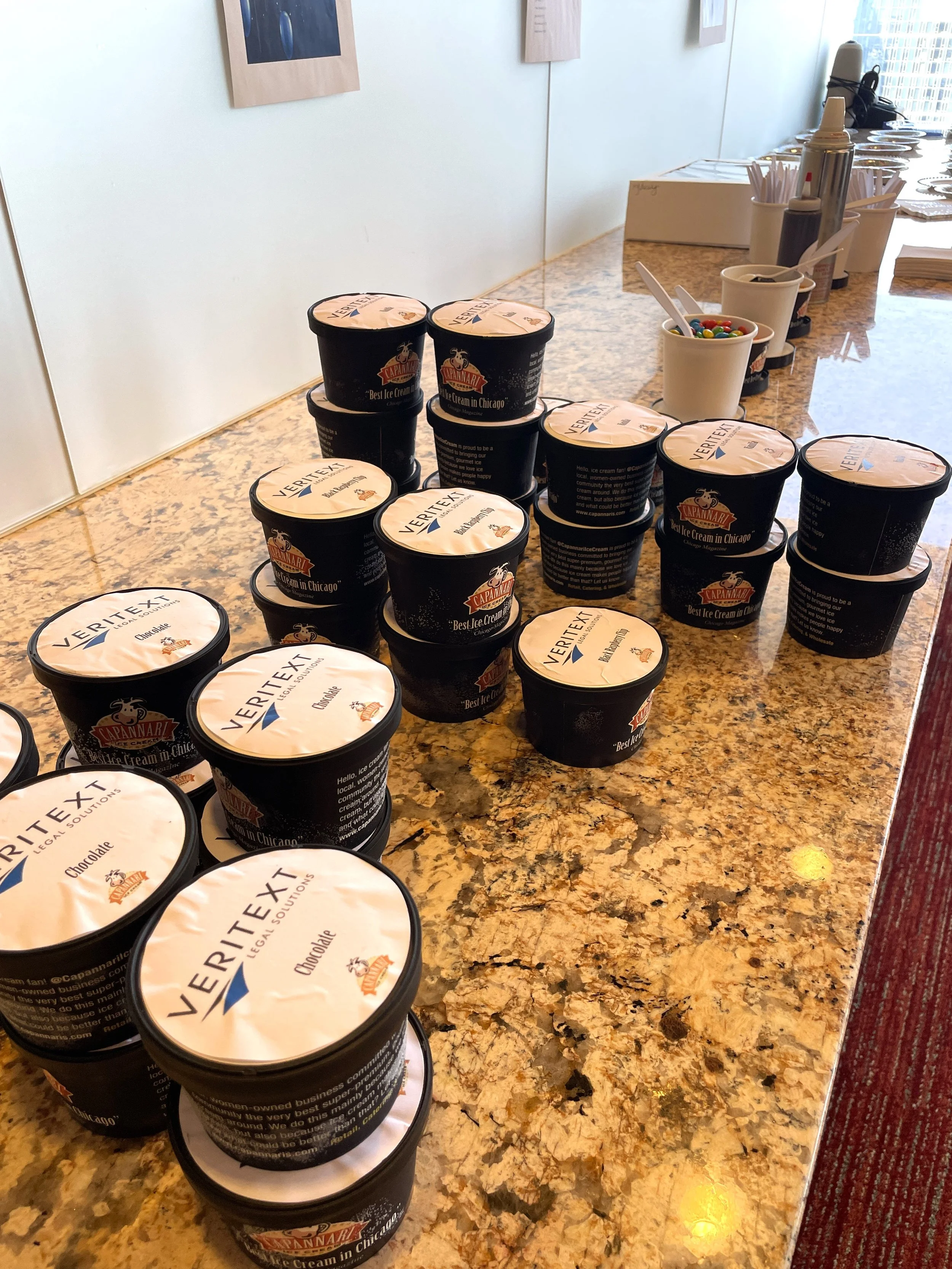 Multiple containers of Capannari ice cream on a granite countertop with cups of toppings and utensils in the background.