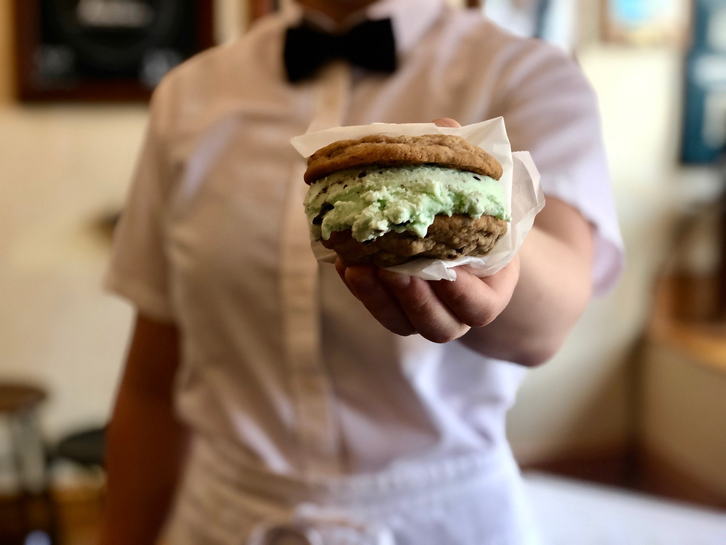 A person in a white shirt with a black bow tie holding a McDonald's ice cream sandwich with a cookie, chocolate chip, and mint flavor, presented in a napkin.