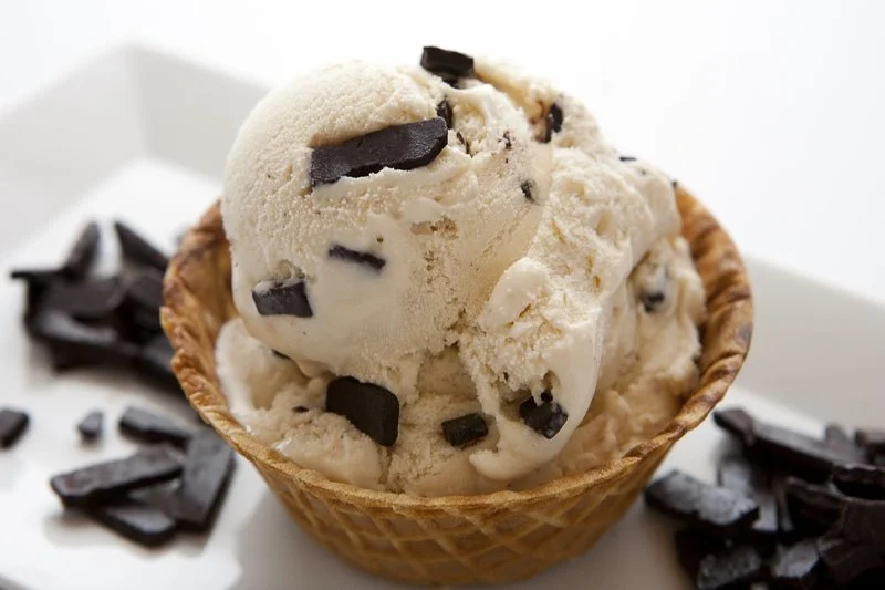 A scoop of vanilla ice cream with chocolate chunks in a waffle cone, surrounded by additional chocolate pieces on a white plate.