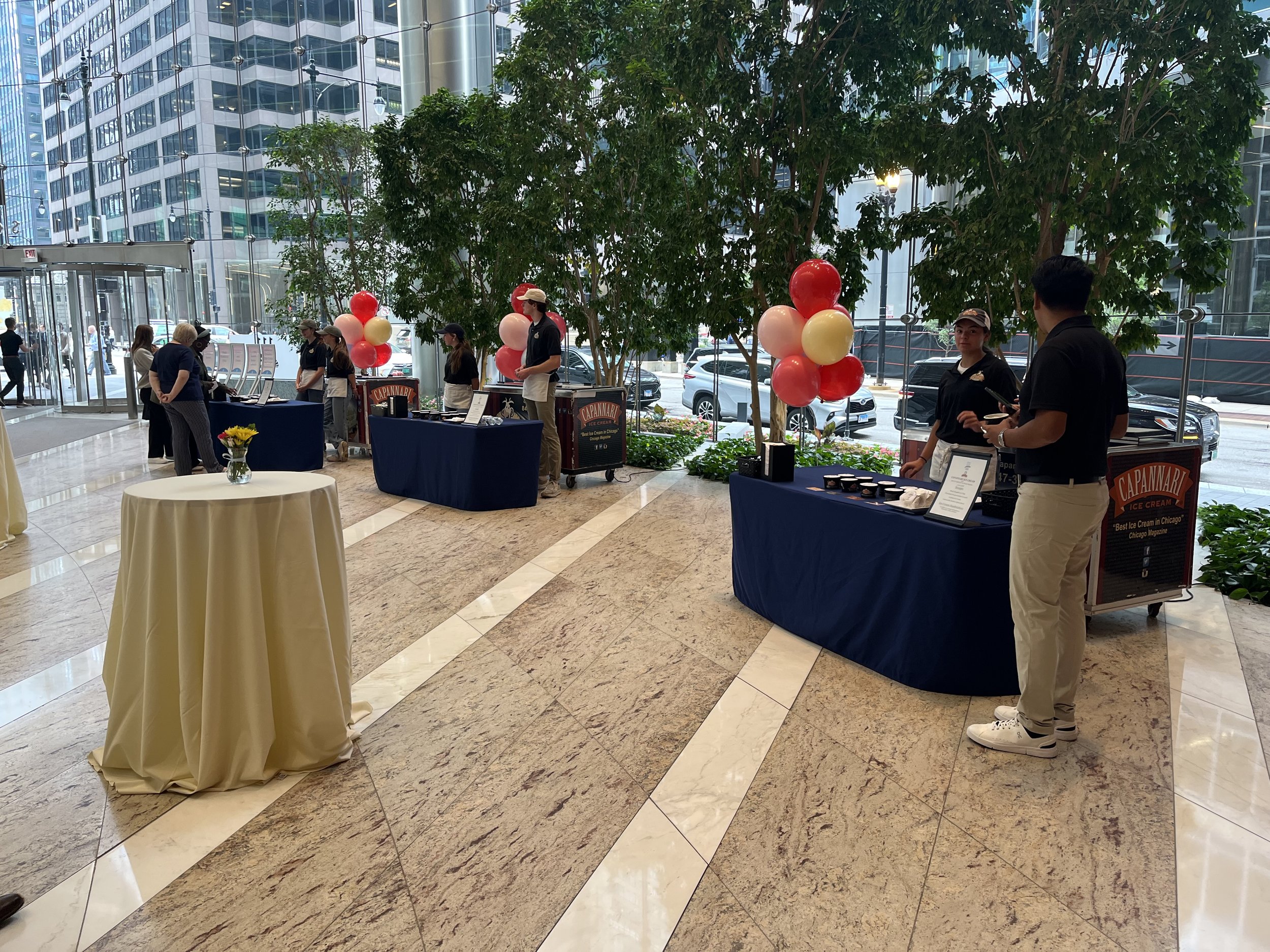 Event setup inside a modern building with several tables, balloons, and staff attending to visitors. A table with a vase of yellow flowers is in the foreground, large indoor trees, and high-rise buildings visible outside through glass walls.