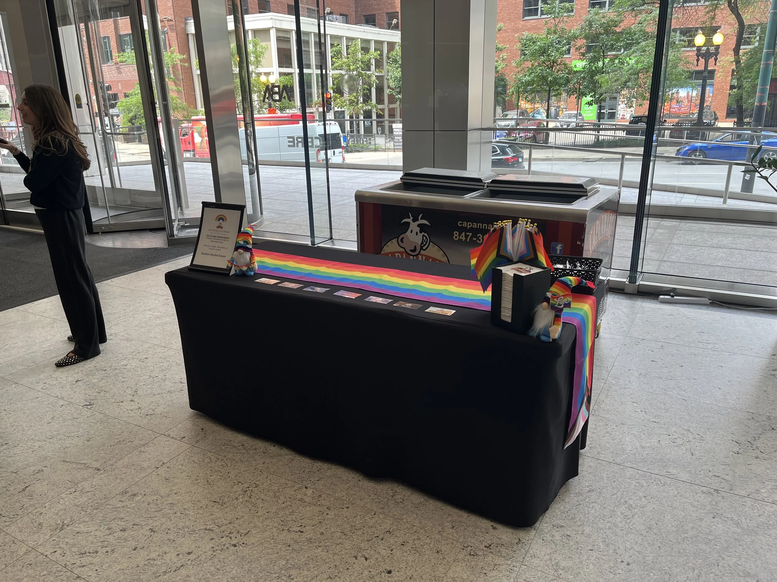 Table with LGBT pride rainbow flags, rainbow-colored cloth, rainbow-themed plush toys, pamphlets, and signs inside a building lobby near glass doors and large windows overlooking a city street.