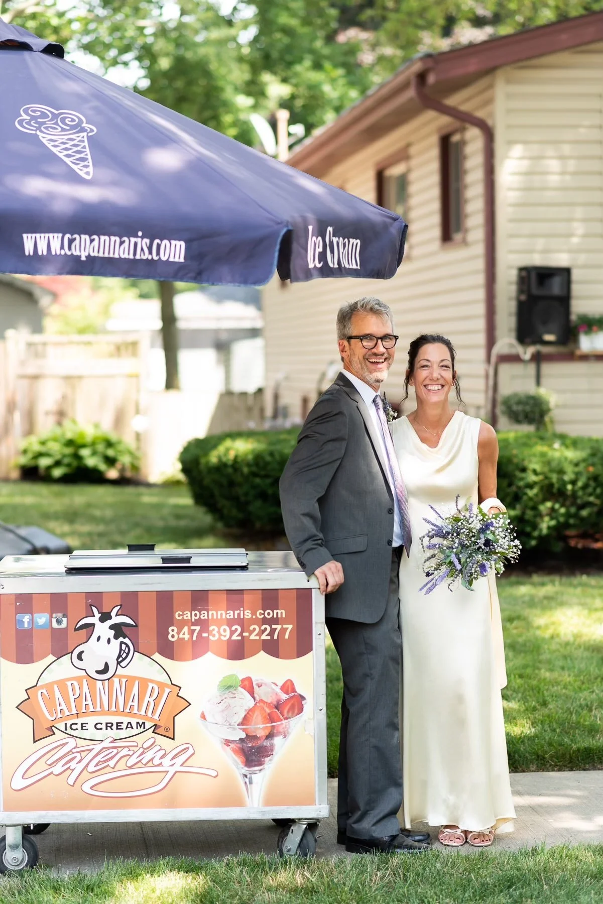 A smiling bride and groom stand outside next to a Capanaris Ice Cream Catering cart with an umbrella overhead.