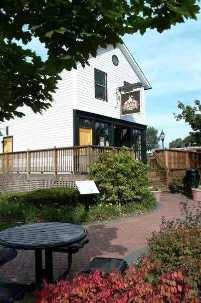 A white two-story building with black trim, a porch with wooden railing, and a sign hanging on the front. There is a tree with green leaves in the foreground, a brick pathway, and outdoor seating with a table and chairs. Bushes and flowers are near the pathway.