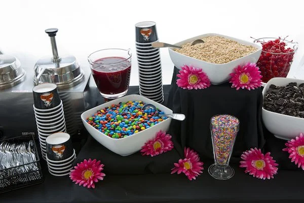A dessert table with bowls of candies, a container of cereal, and bowls of cherries and chocolate chips, decorated with pink flowers.