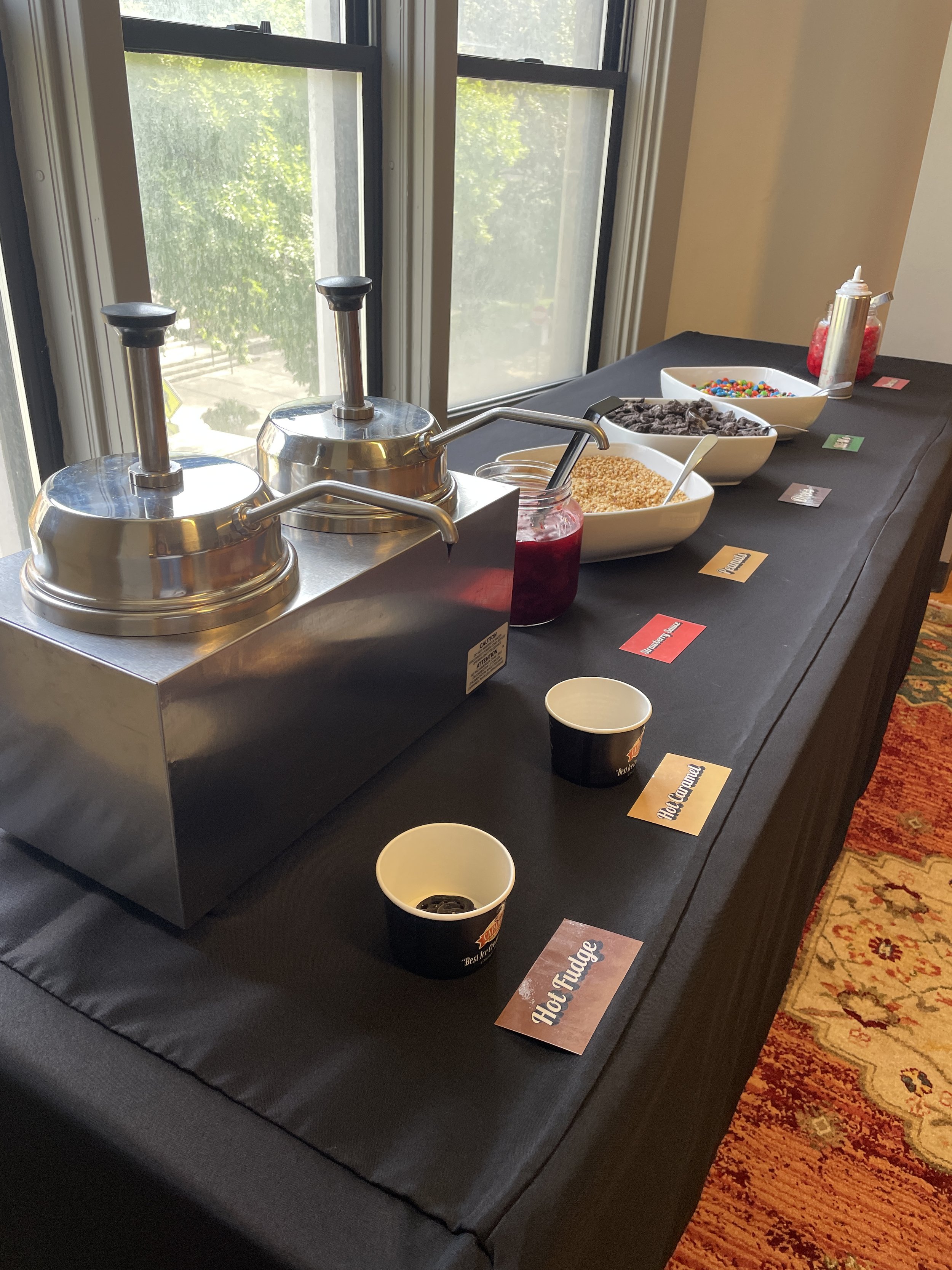 A dessert station with two heated dispensers, bowls of toppings including chocolate chips, sprinkles, and crushed nuts, and small cups for serving. The setup is next to a window with a view of trees outside.