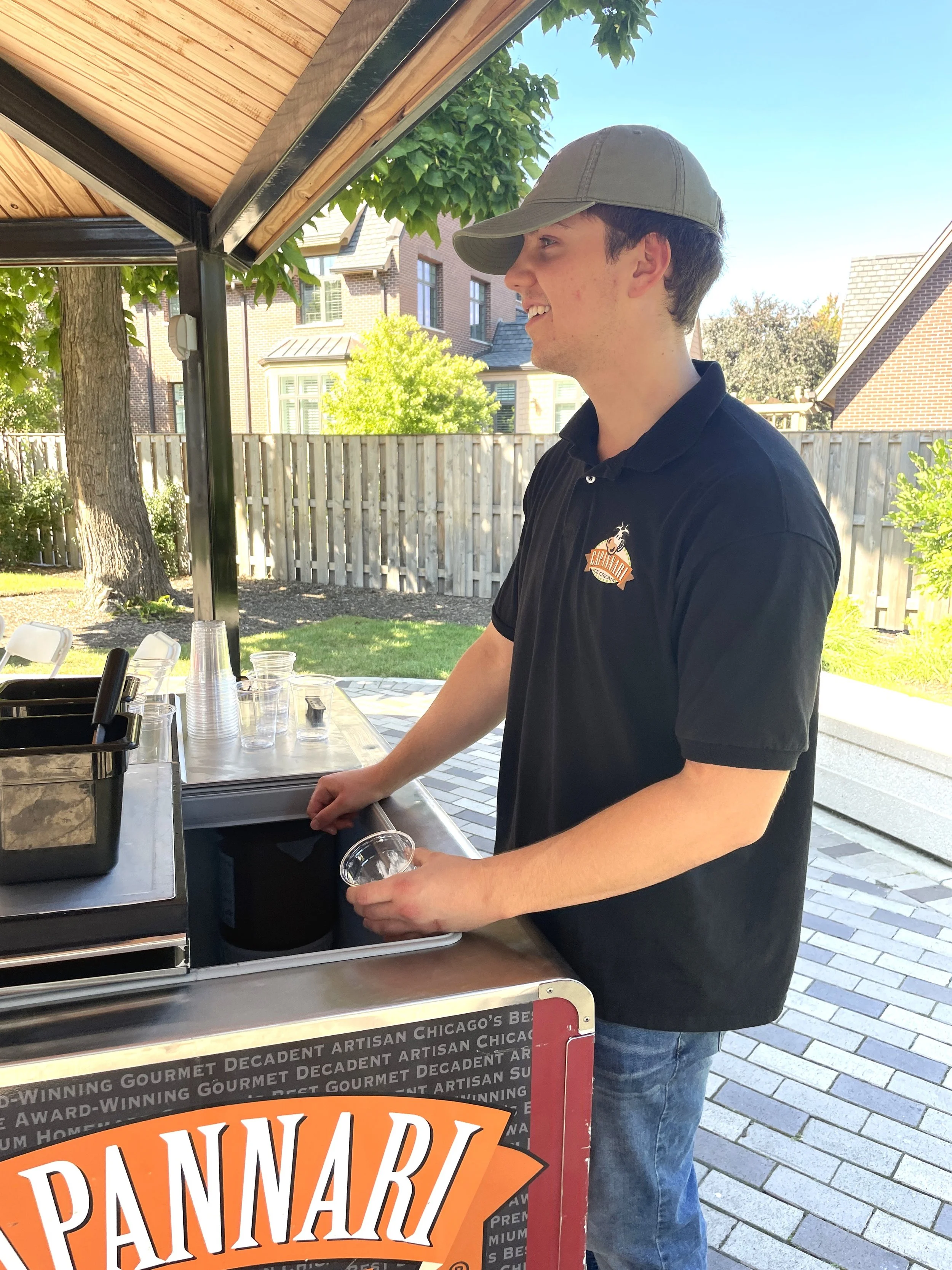 A young man wearing a gray cap and black polo shirt serving ice cream at an outdoor food stand with a wooden roof, with a background of trees, a wooden fence, and residential houses.
