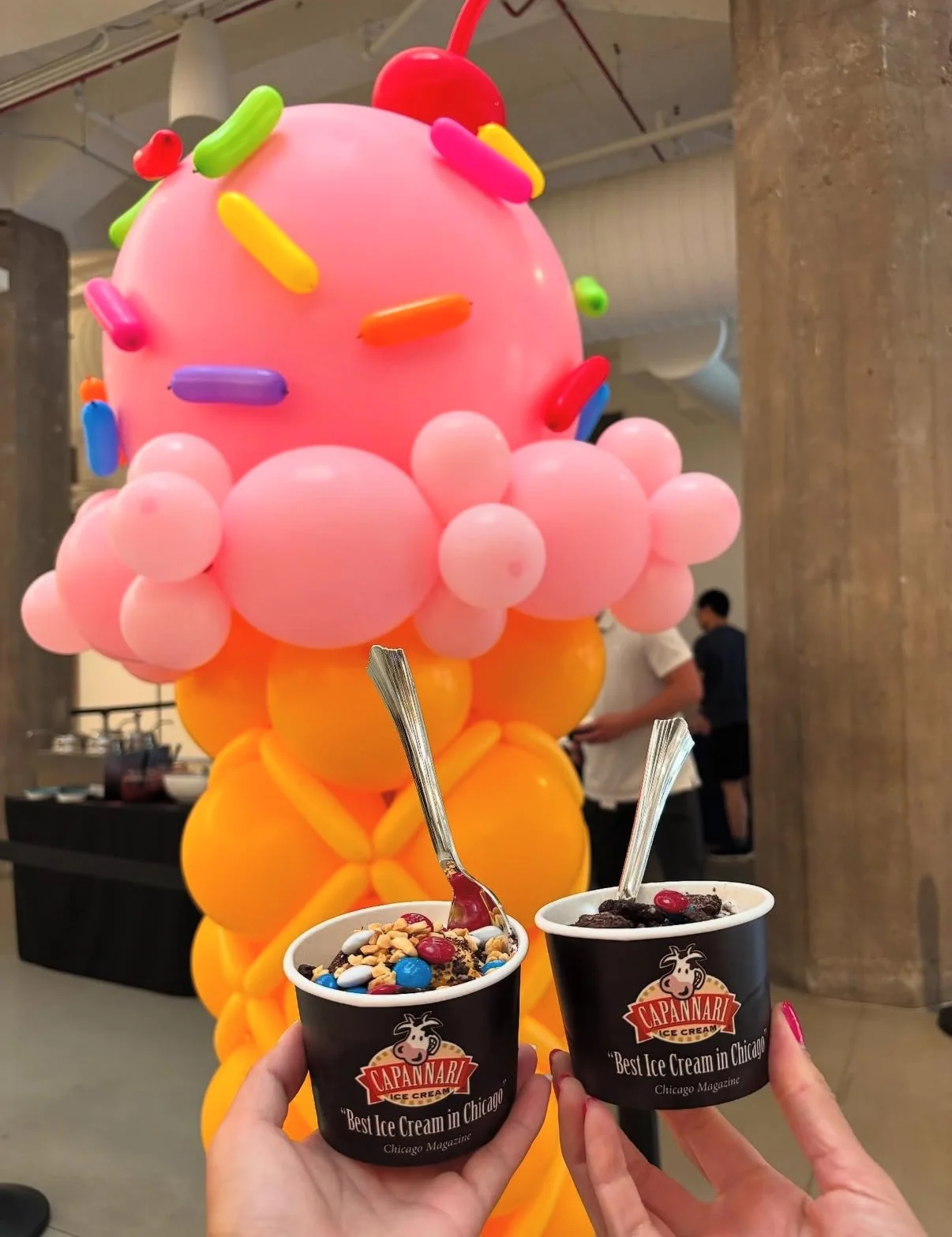 Two cups of ice cream in black cups with the Capannari logo, held by two hands in front of a colorful balloon sculpture resembling an ice cream cone.