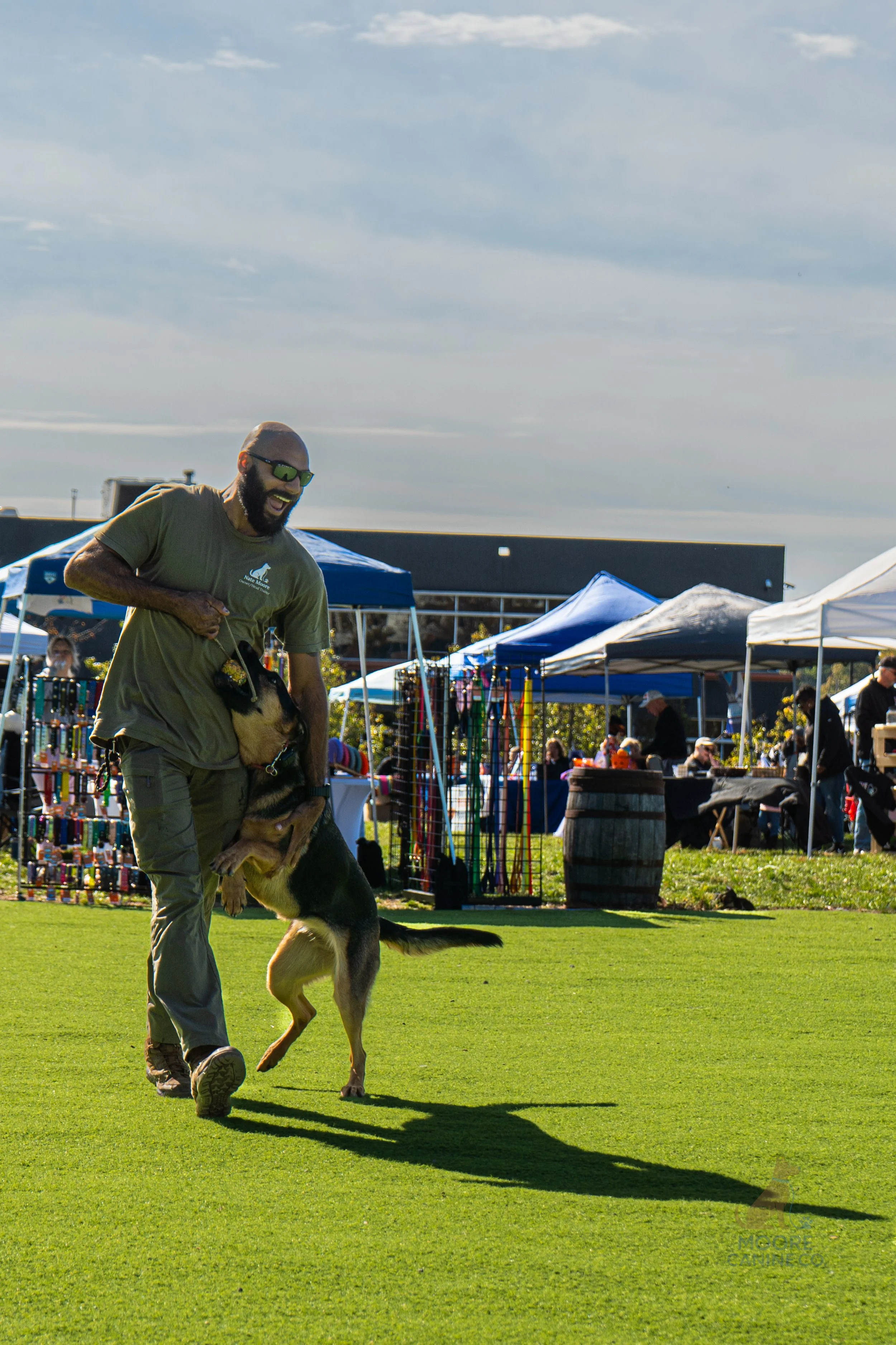 A man and a German Shepherd playing tug-of-war during an outdoor event with vendor tents and people in the background.