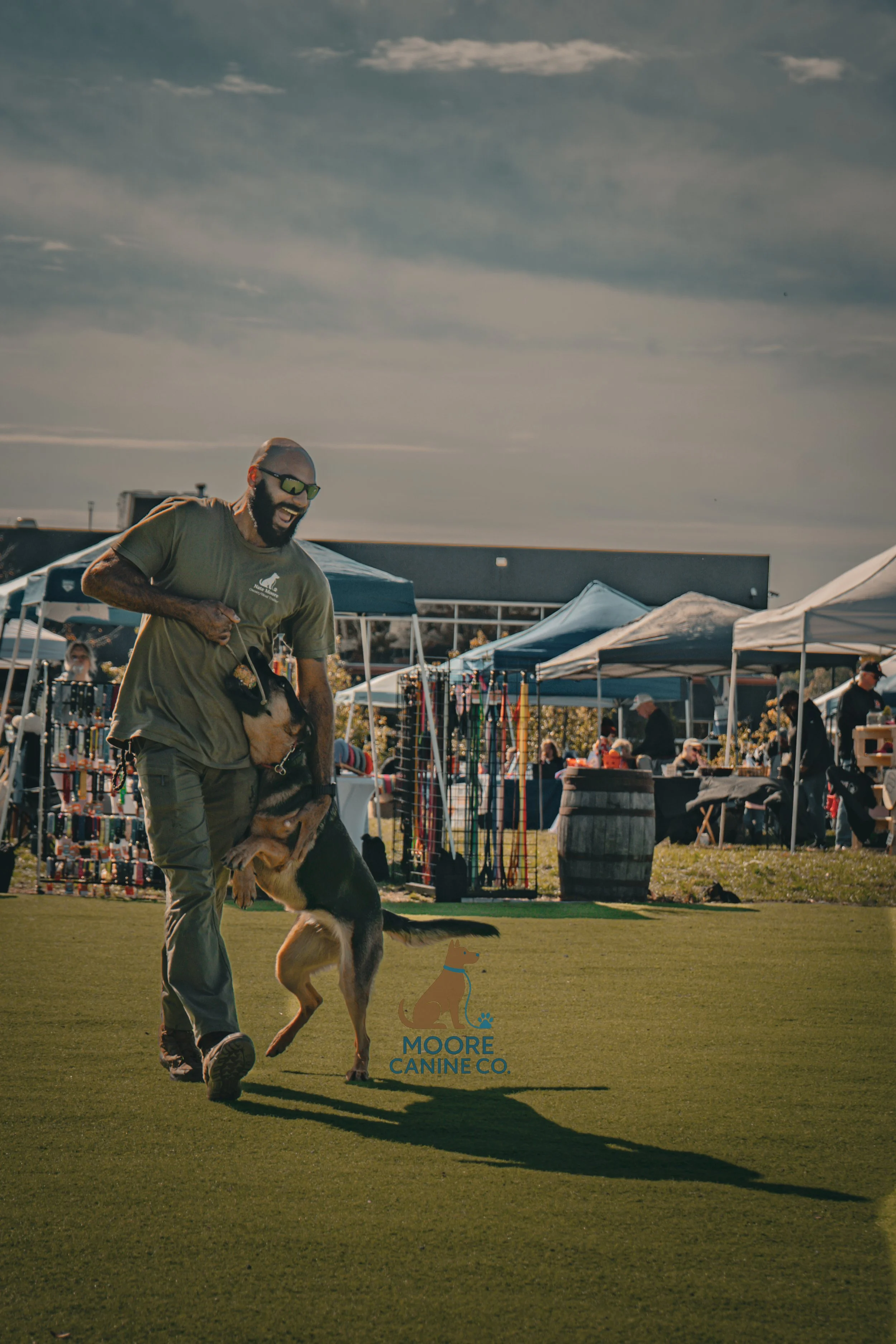 A man with sunglasses and a beard, wearing a green t-shirt and cargo pants, is smiling and playing with a German Shepherd dog at an outdoor event with vendor tents and people in the background.