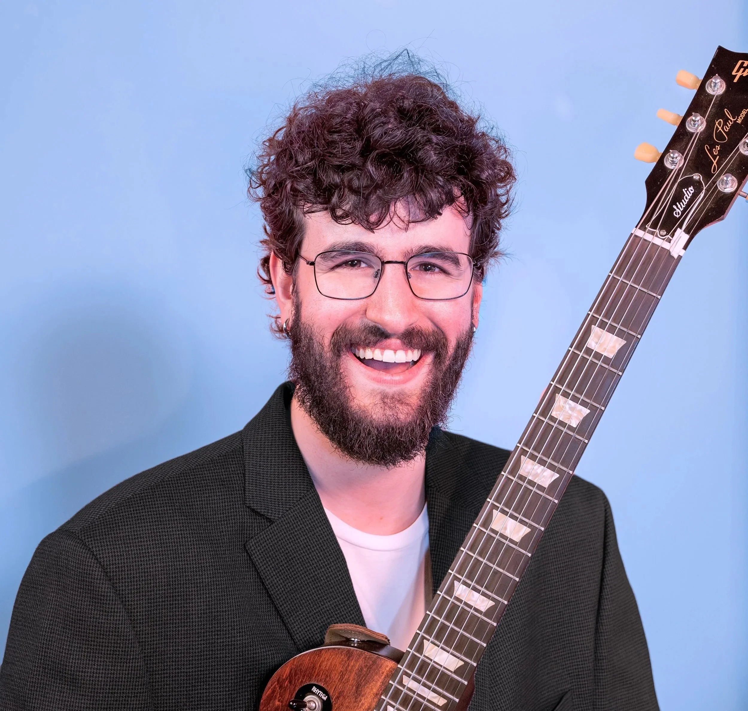 Close-up of a smiling man with curly brown hair and glasses, holding a guitar against a light blue background.