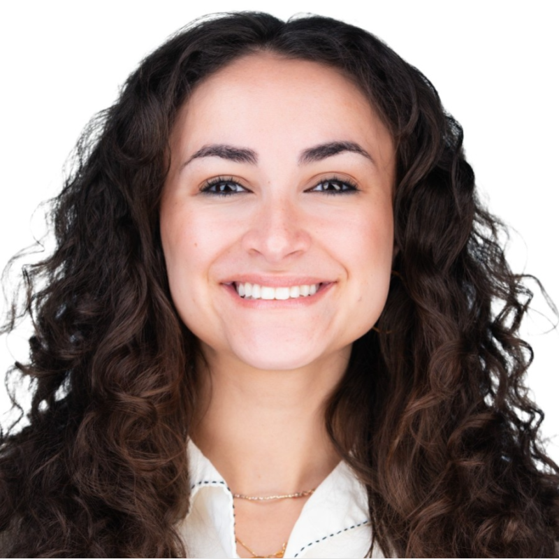 Portrait of a smiling young woman with curly brown hair, wearing a white blouse and a gold necklace, against a white background.