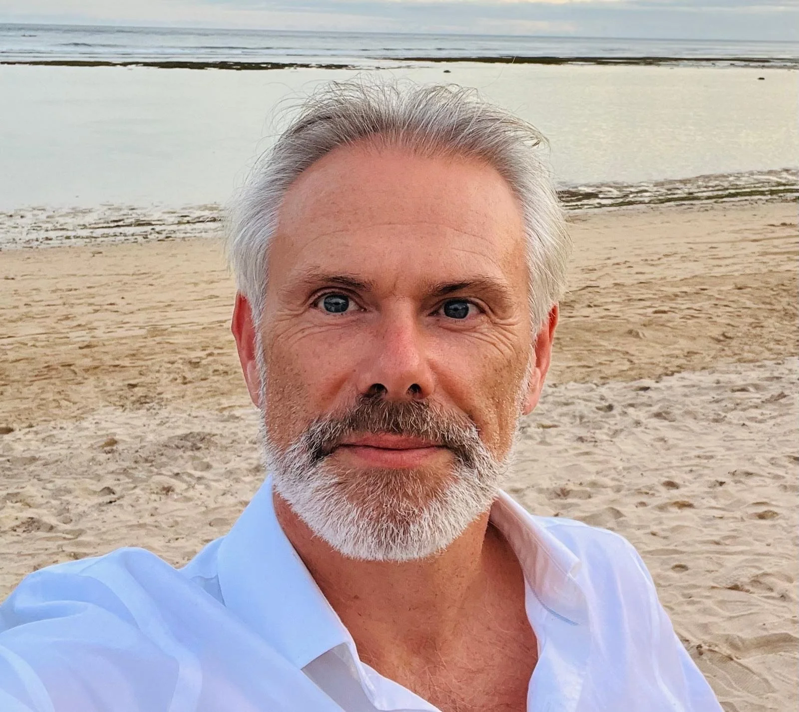 A man with gray hair and beard taking a selfie on a sandy beach with water and a cloudy sky in the background.