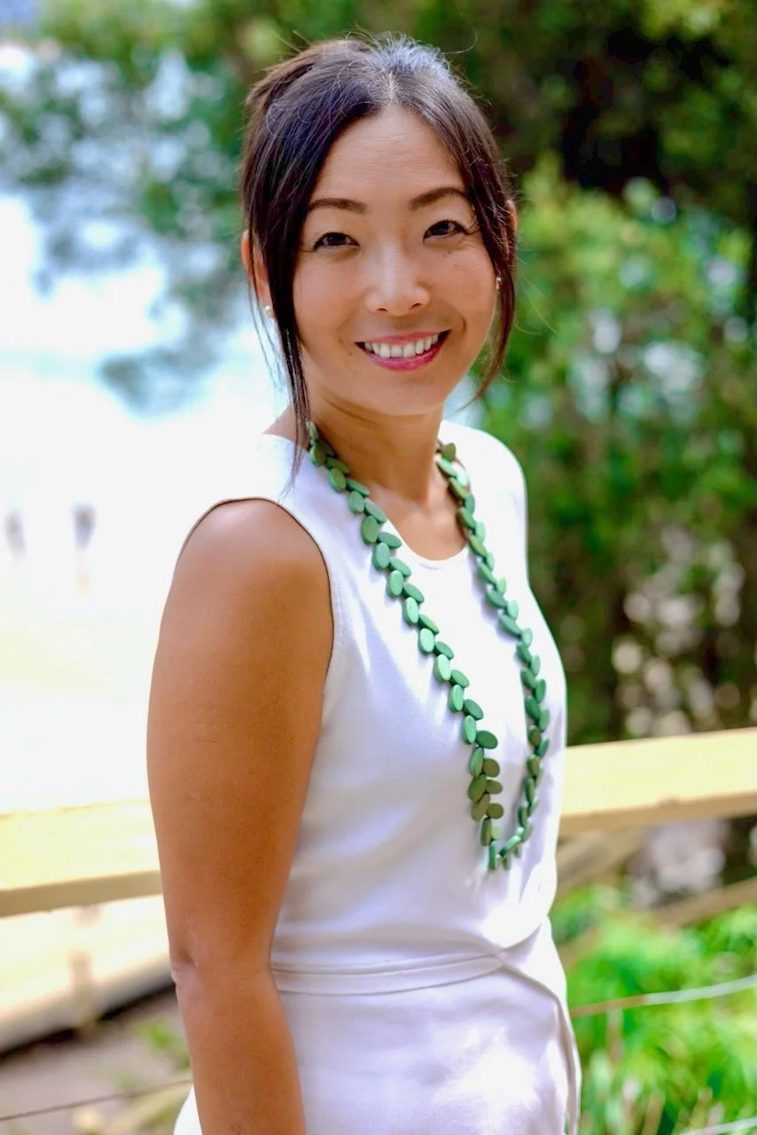 A smiling Japanese Australian woman in a sleeveless white top with a green beaded necklace outdoors with trees and greenery in the background. She is an expert in wellness and sound healing.