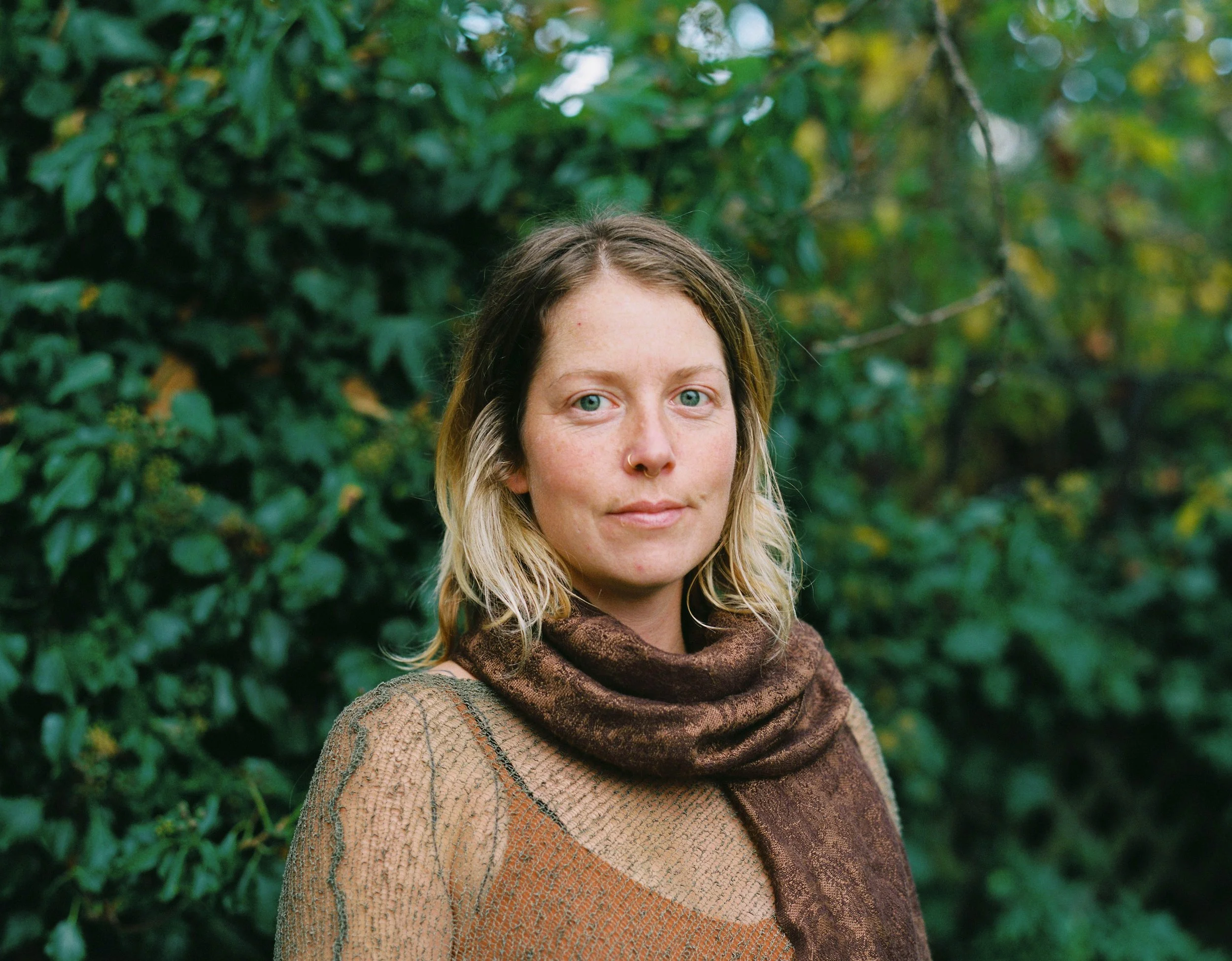 A woman with shoulder-length blonde hair and blue eyes standing outdoors in front of green foliage, wearing a light brown sweater and a dark brown scarf.