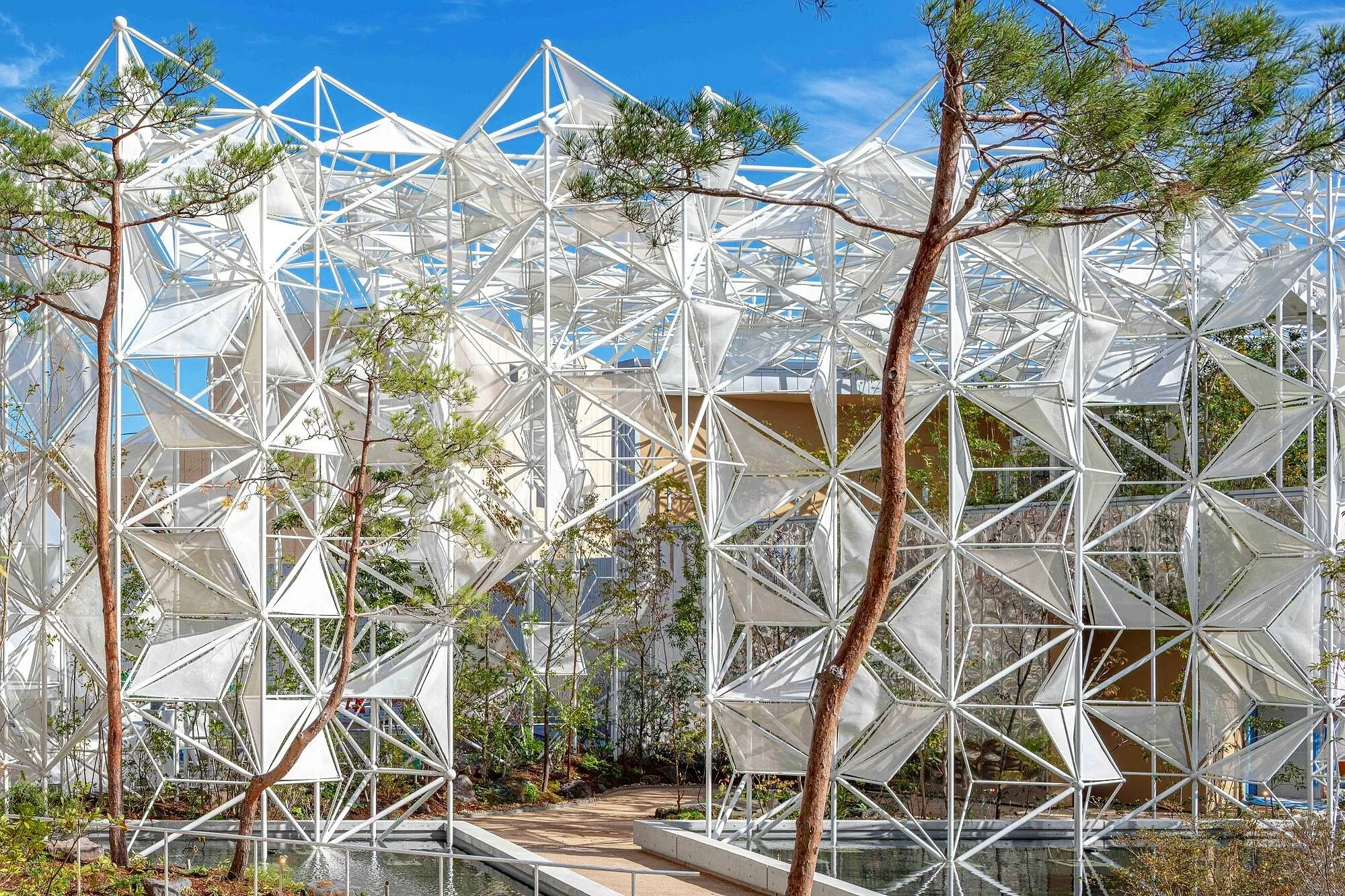 Modern architectural structure with a white geometric framework, surrounded by trees and a small pond, under a bright blue sky.