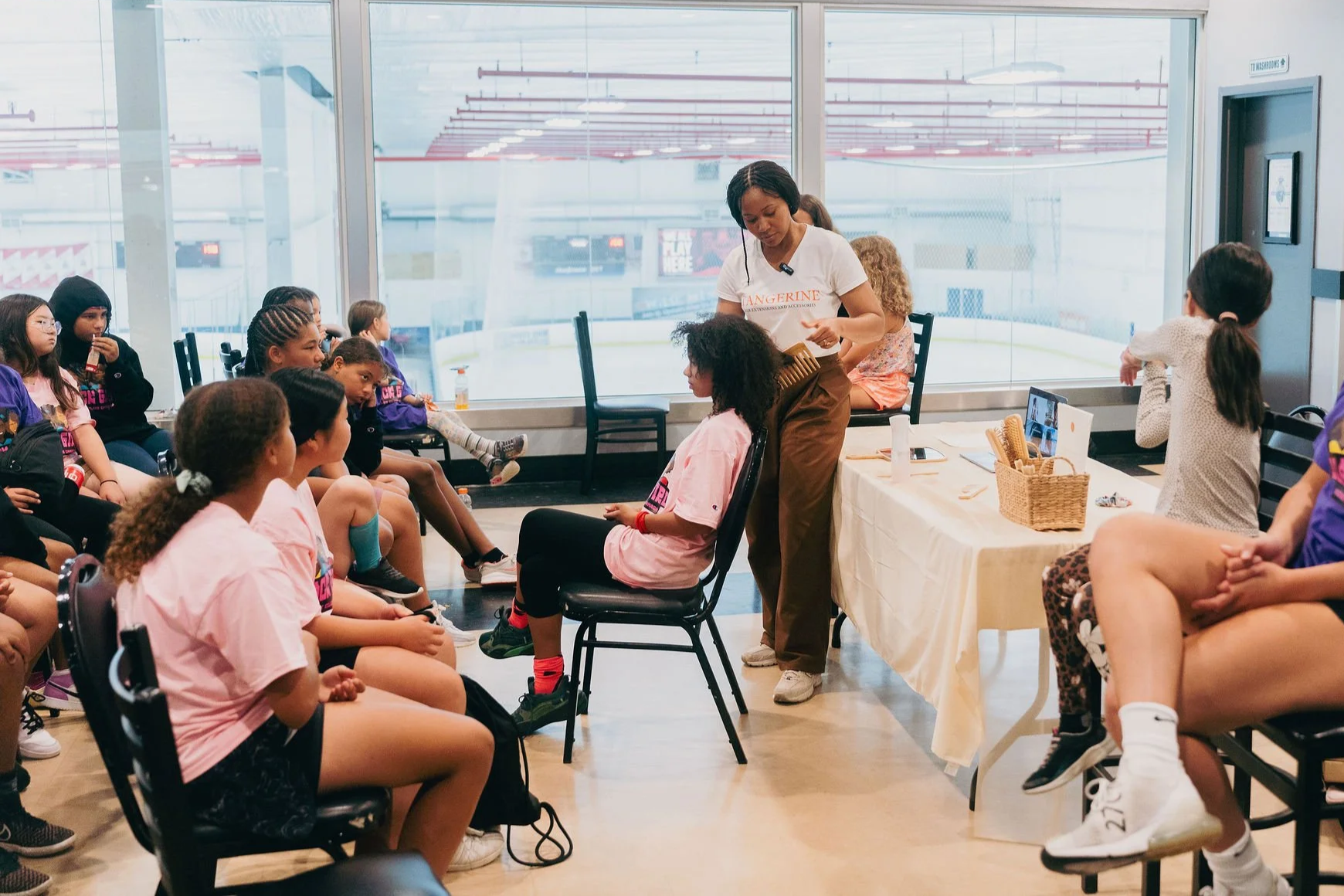 A group of children attending a workshop or class at an indoor ice rink facility. The instructor, a woman, is demonstrating something to a seated girl while others watch. The scene includes a table with supplies and a large window overlooking the ice