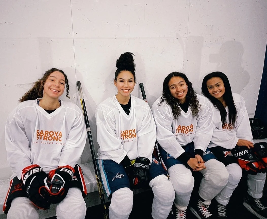 Four young women wearing hockey gear sitting on a bench against a white wall, smiling at the camera.
