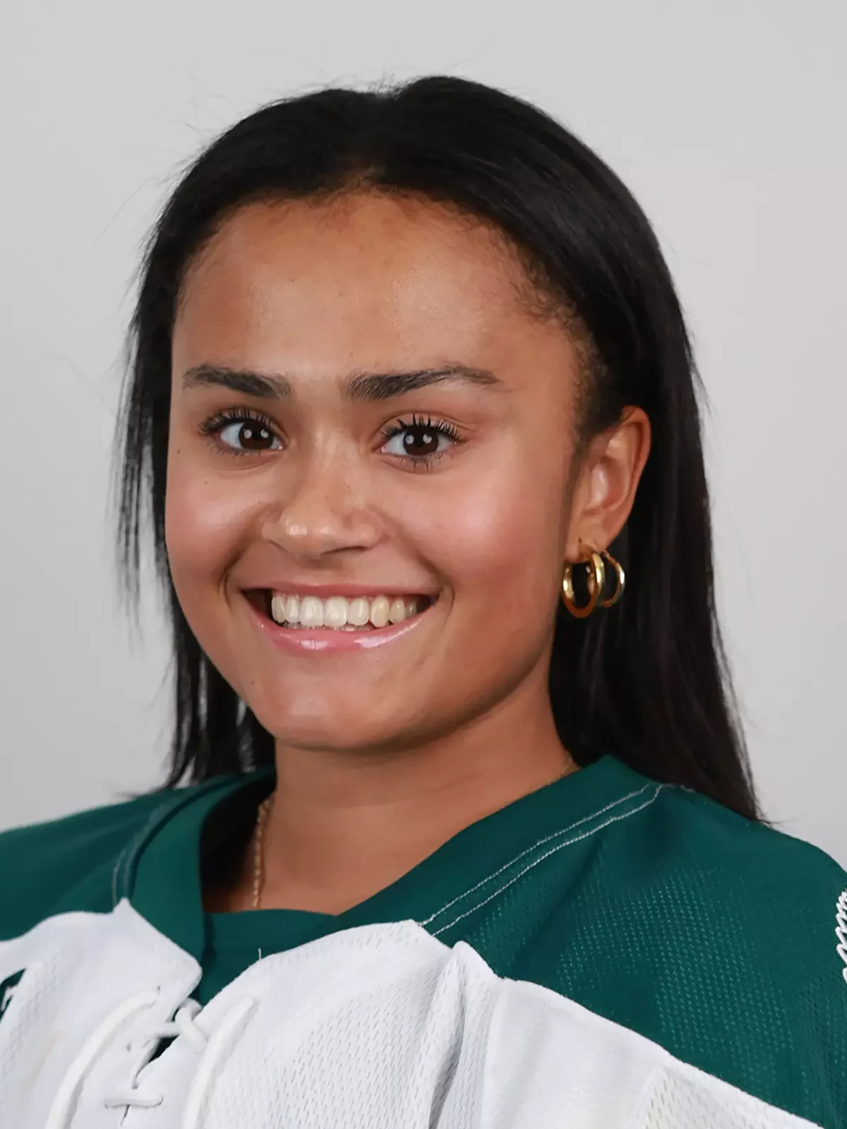 Close-up of a smiling young woman with dark hair, gold hoop earrings, and wearing a green and white sports jersey.