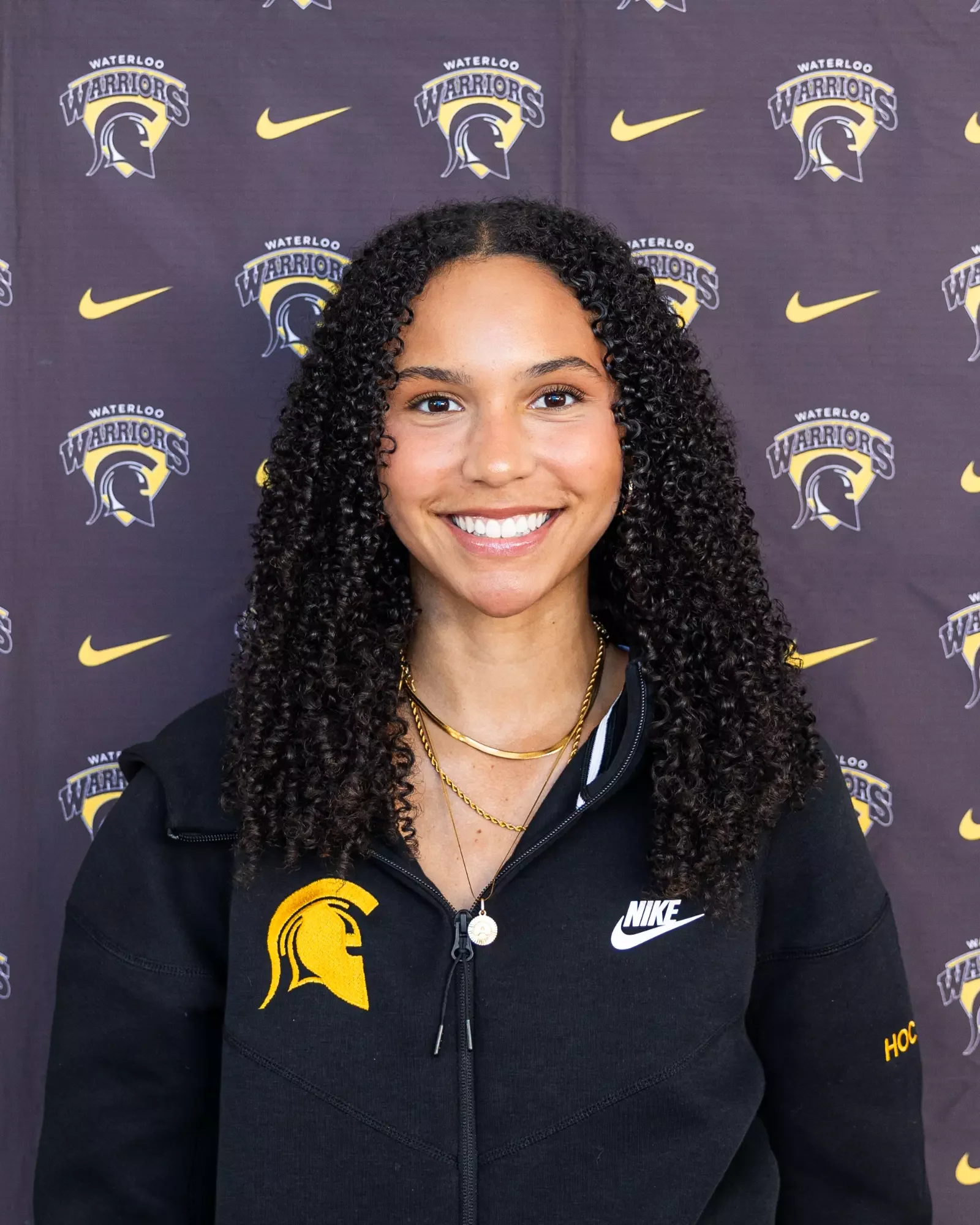 Young woman with curly hair smiling, wearing a black Nike hoodie with Waterloo Warriors logo, standing in front of a backdrop with Waterloo Warriors logos and Nike swooshes.