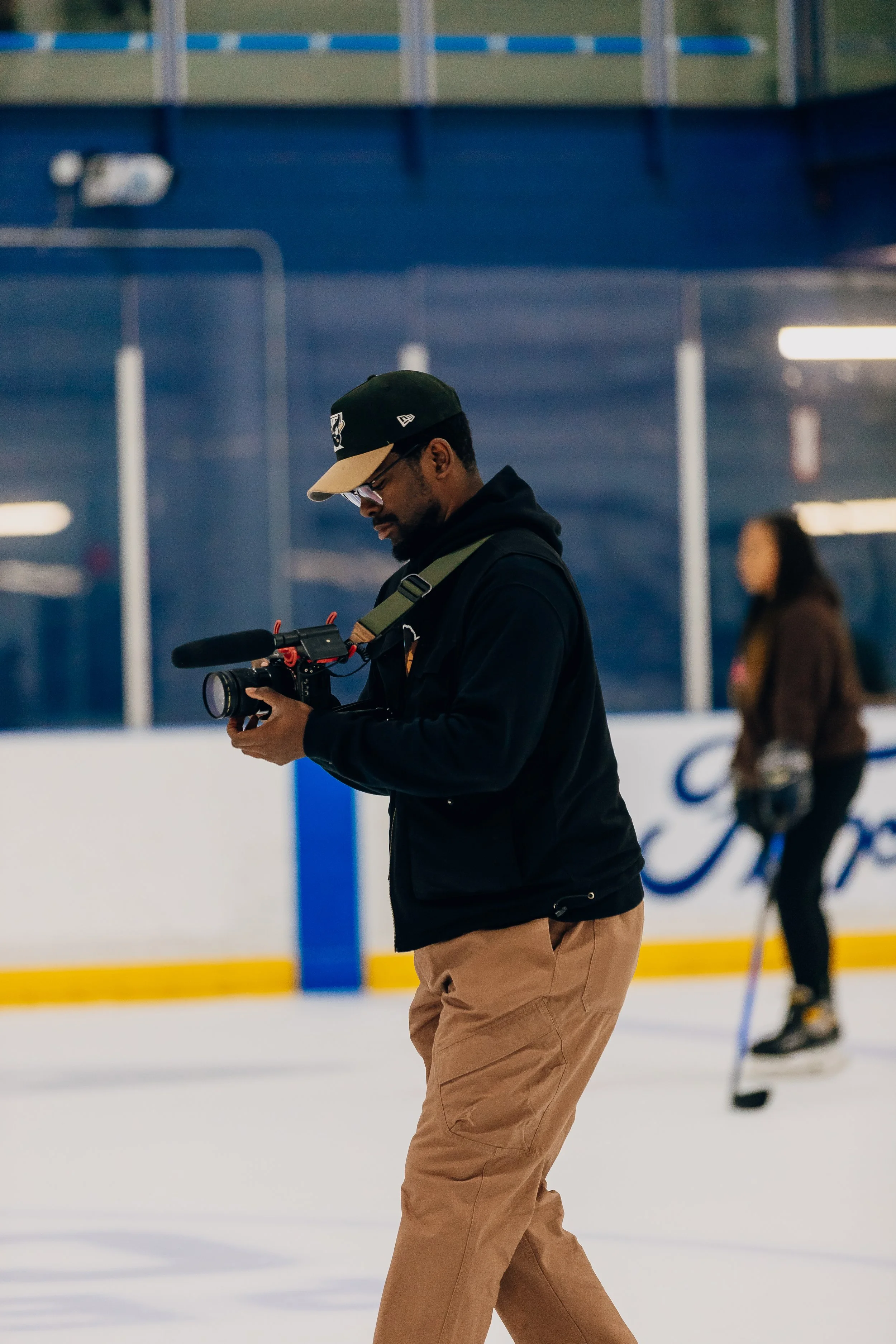 A man in a black jacket and tan pants walks on an ice rink, looking at a camera he is holding, with a woman skating in the background.