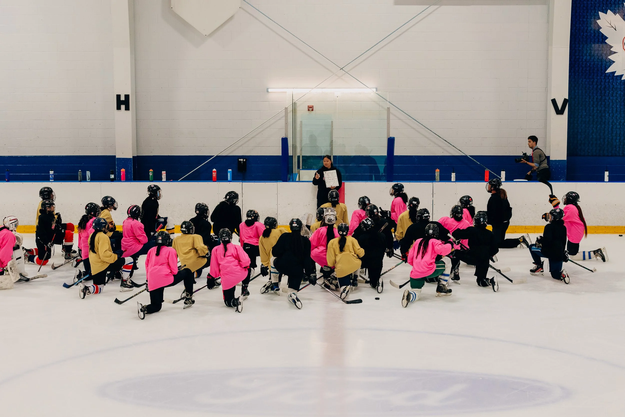 A group of children wearing hockey gear, including helmets and skates, are gathered on an ice rink listening to a coach or instructor. The kids are dressed in black, pink, yellow, and other colors. An adult is standing in front of them holding a board, and another person is to the right holding a camera or recording device.