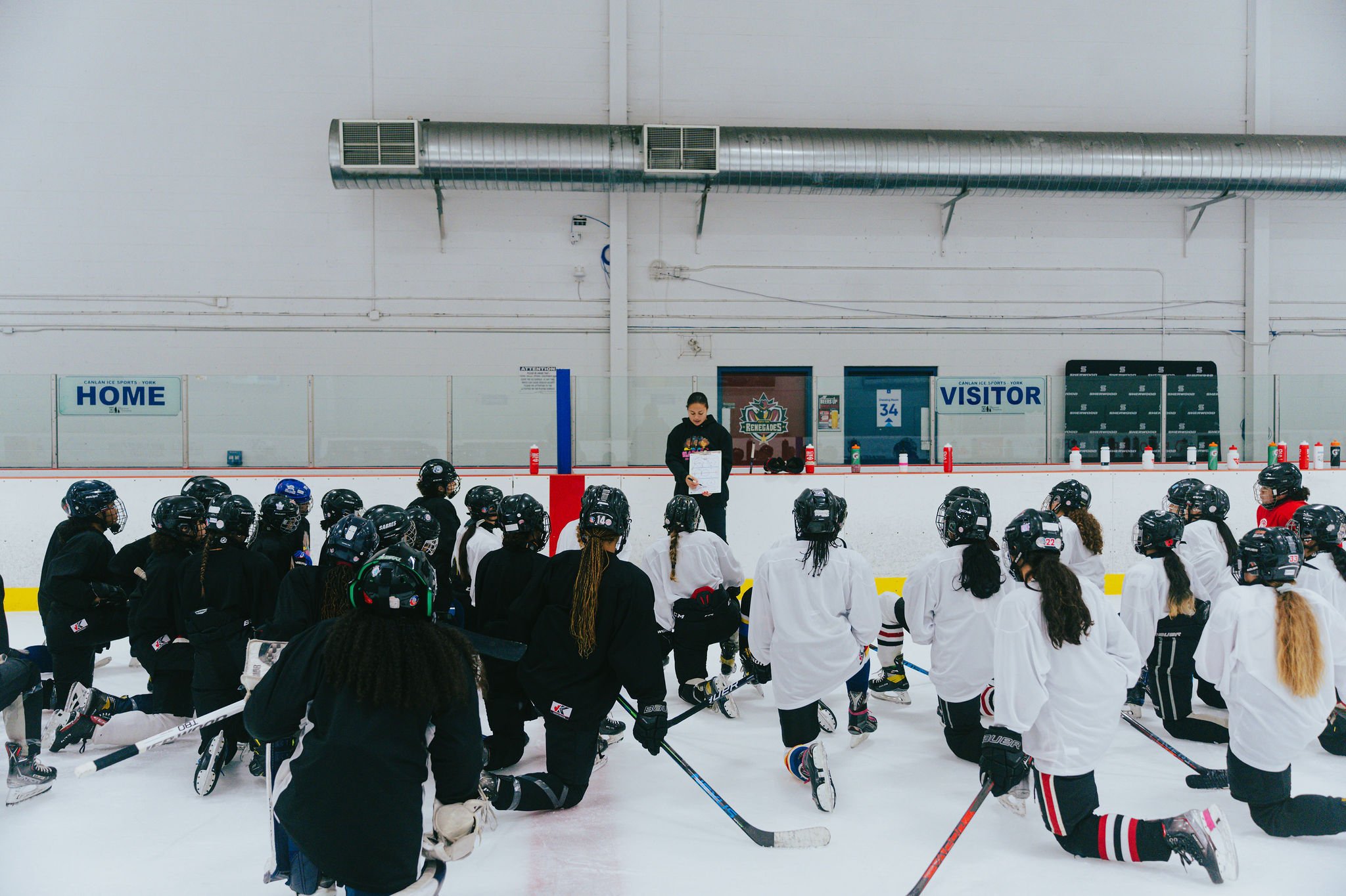 A group of female hockey players, some kneeling, listening to a coach on the ice rink. The coach is standing in front of the players holding a clipboard. The rink has a white wall with signs indicating "Home" and "Visitor."