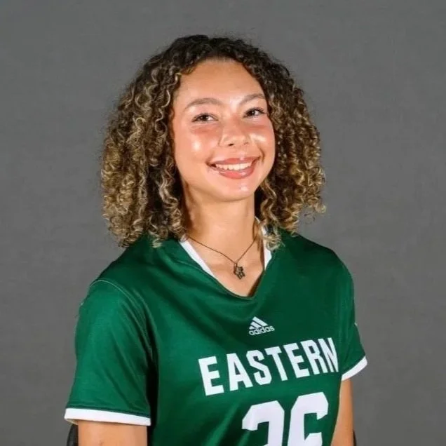 A young woman with curly hair wearing a green sports jersey with the number 26 and 'Eastern' written on it, smiling against a gray background.