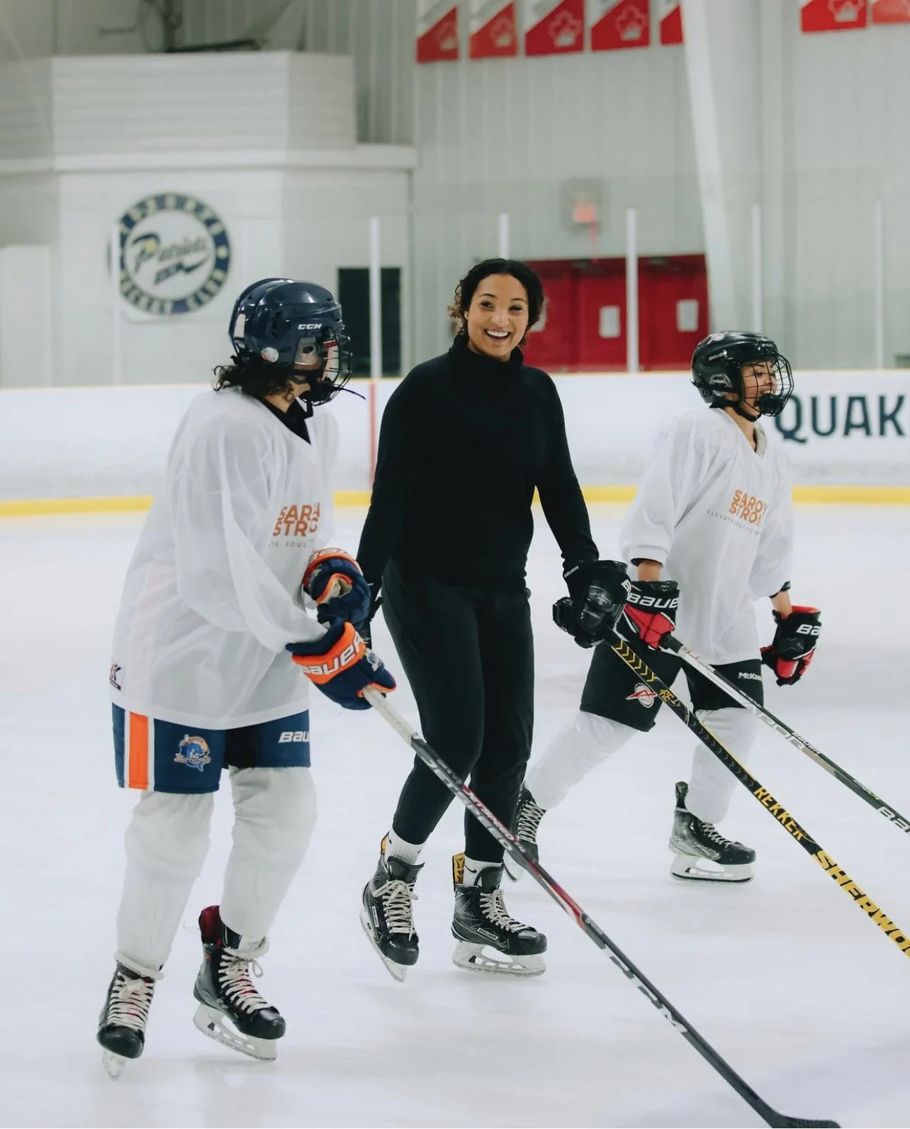 A woman in black skating gear sharing a joyful moment with two children in hockey practice at an indoor ice rink.