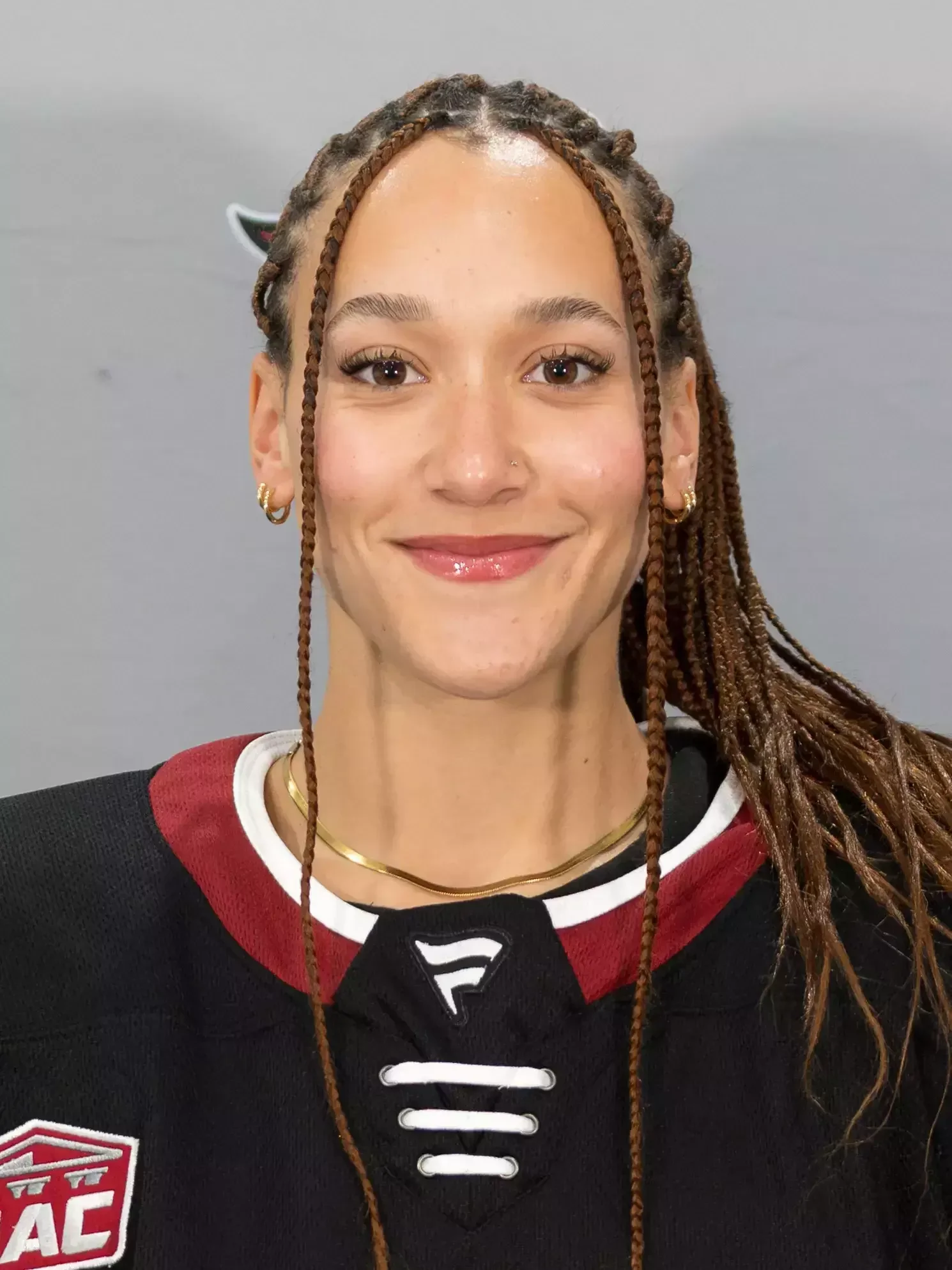 Close-up portrait of a young woman with braided hair, wearing an athletic jersey and jewelry, smiling at the camera.