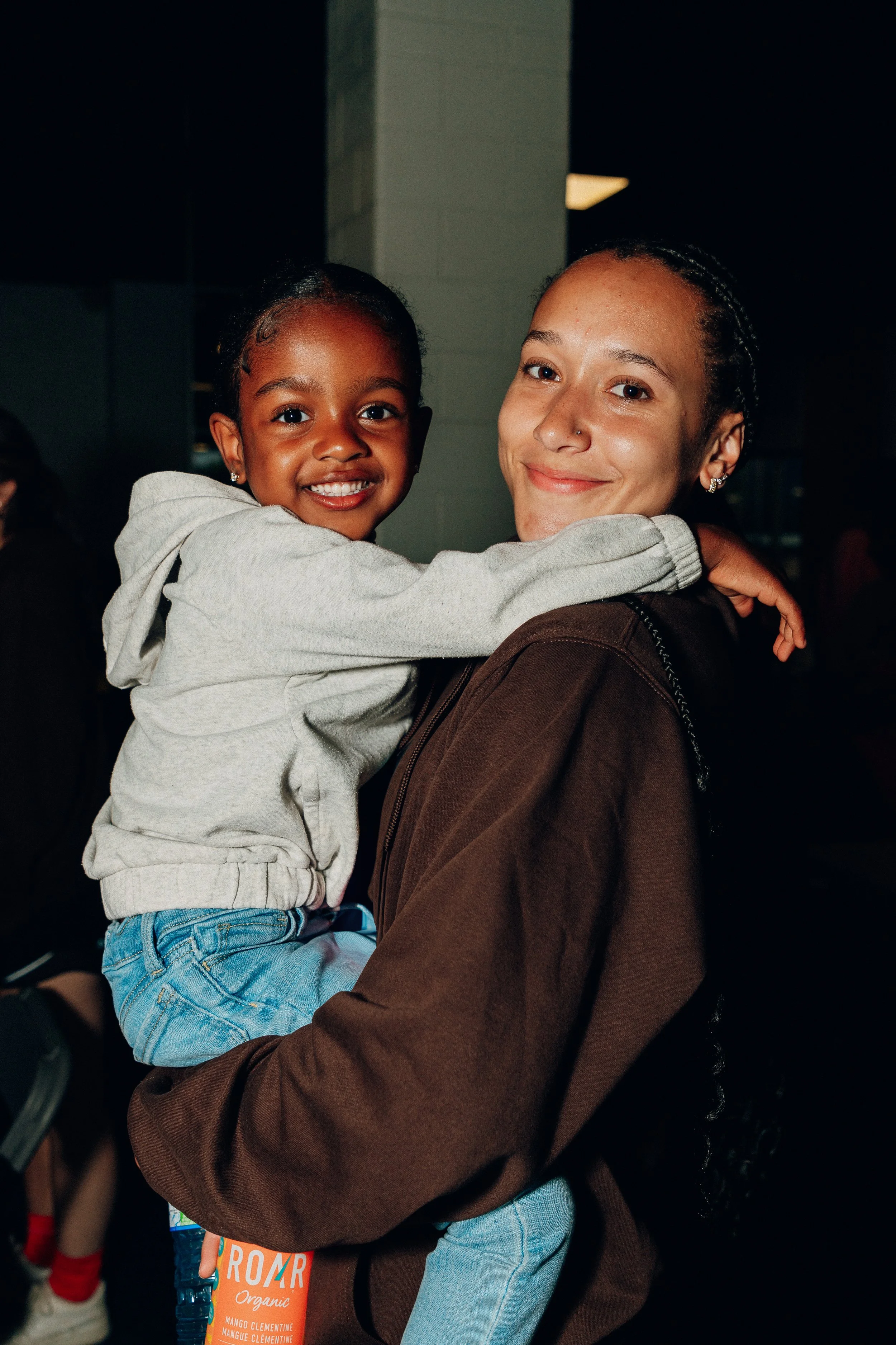 A woman holding a young girl indoors. The girl is smiling and hugging the woman, both looking at the camera.