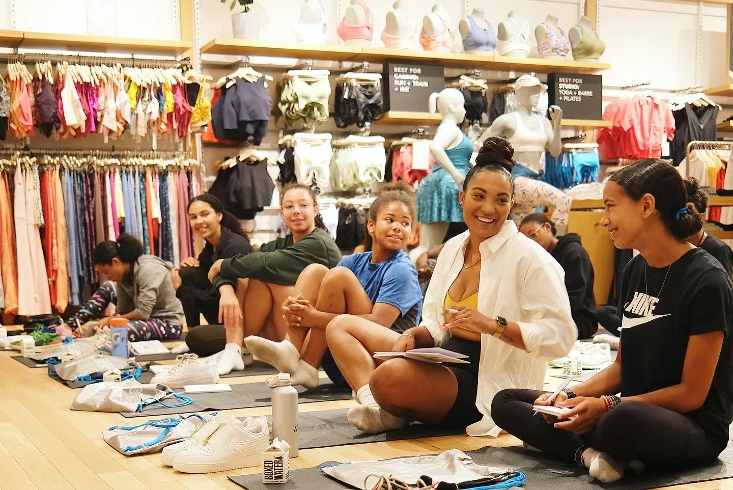 A group of women and girls seated on the floor inside a fitness or dance apparel store, engaged in a discussion or class, with clothing racks and mannequins in the background.