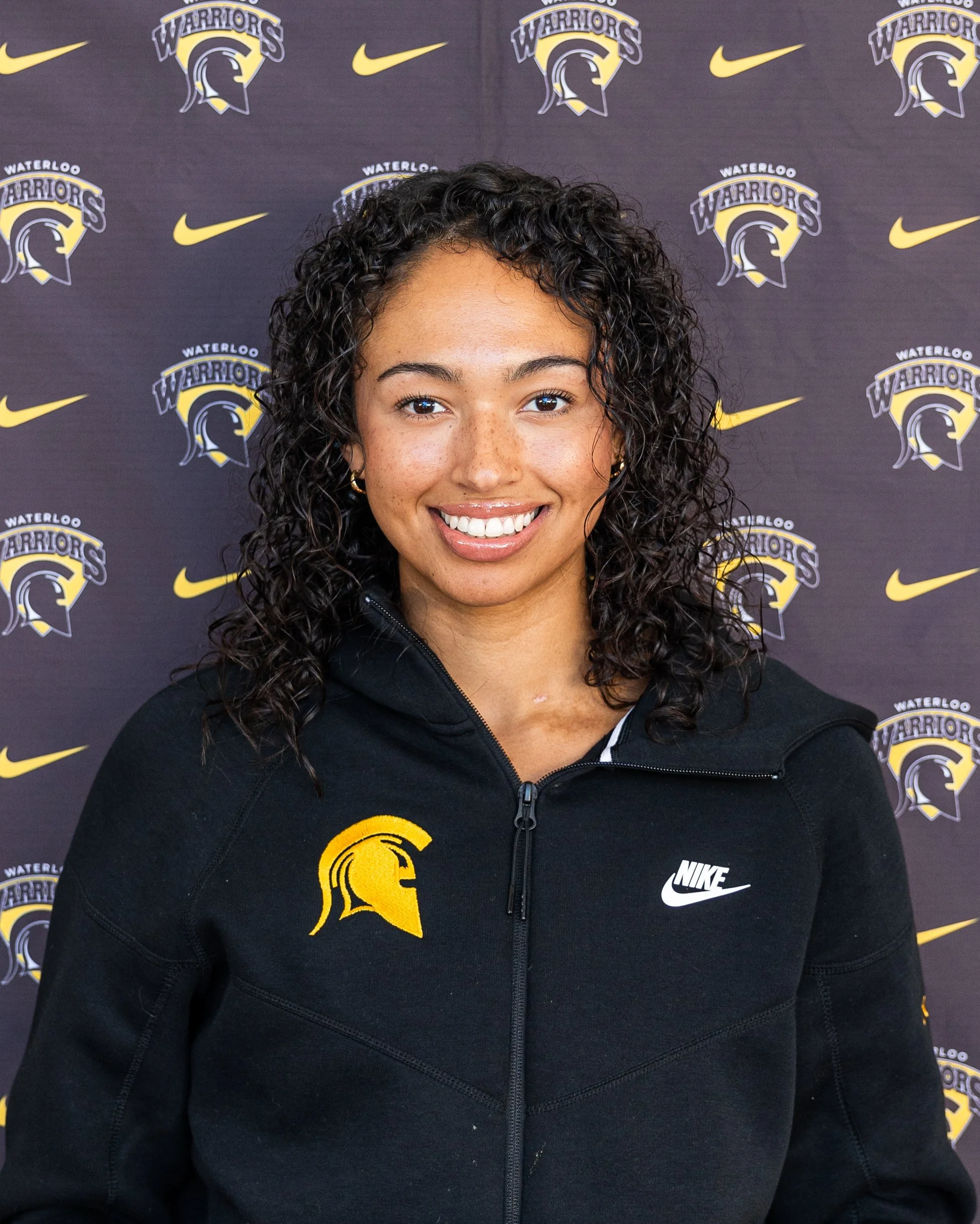 A young woman with curly dark hair and a big smile, wearing a black Nike jacket with a yellow Sparta helmet logo, standing in front of a backdrop with Waterloo Warriors logos and Nike swoosks.