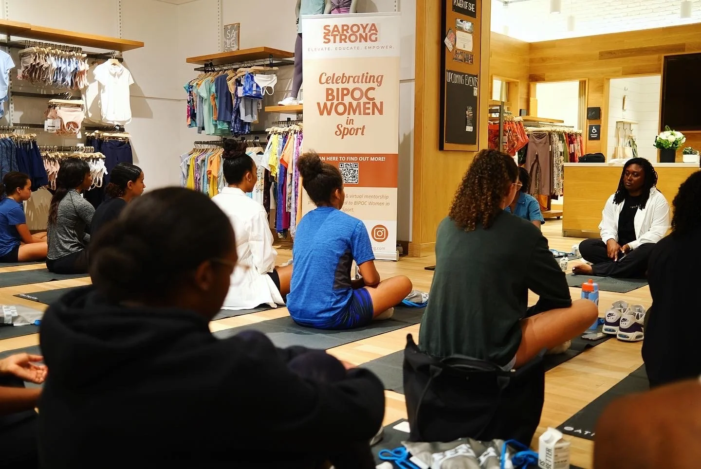 Group of women and girls sitting on yoga mats in a clothing store for a workshop or discussion. A staff member or instructor is seated in front of them near a banner that reads 'Celebrating BIPOC Women in Sport.' The store has various clothing items on racks and a wooden interior.