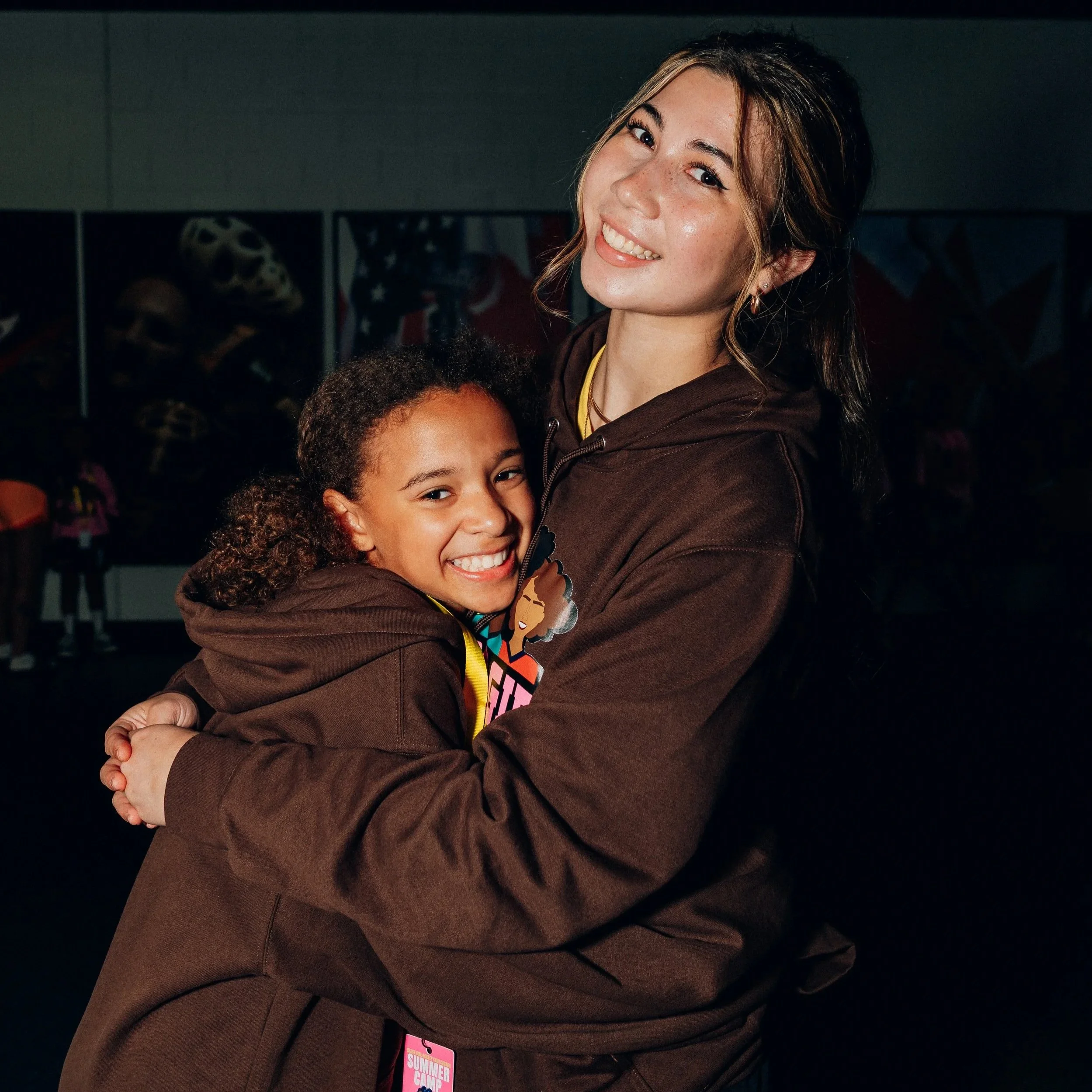 Two girls hugging each other and smiling at the camera, wearing brown hoodies, indoors with some artwork and people in the background.