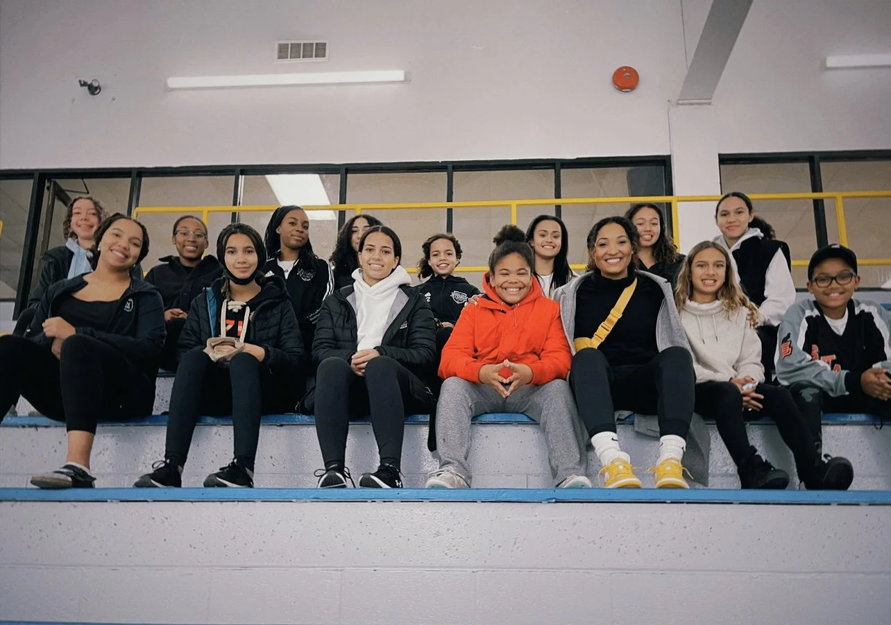 Group of young girls sitting on bleachers in a gymnasium, smiling at the camera.