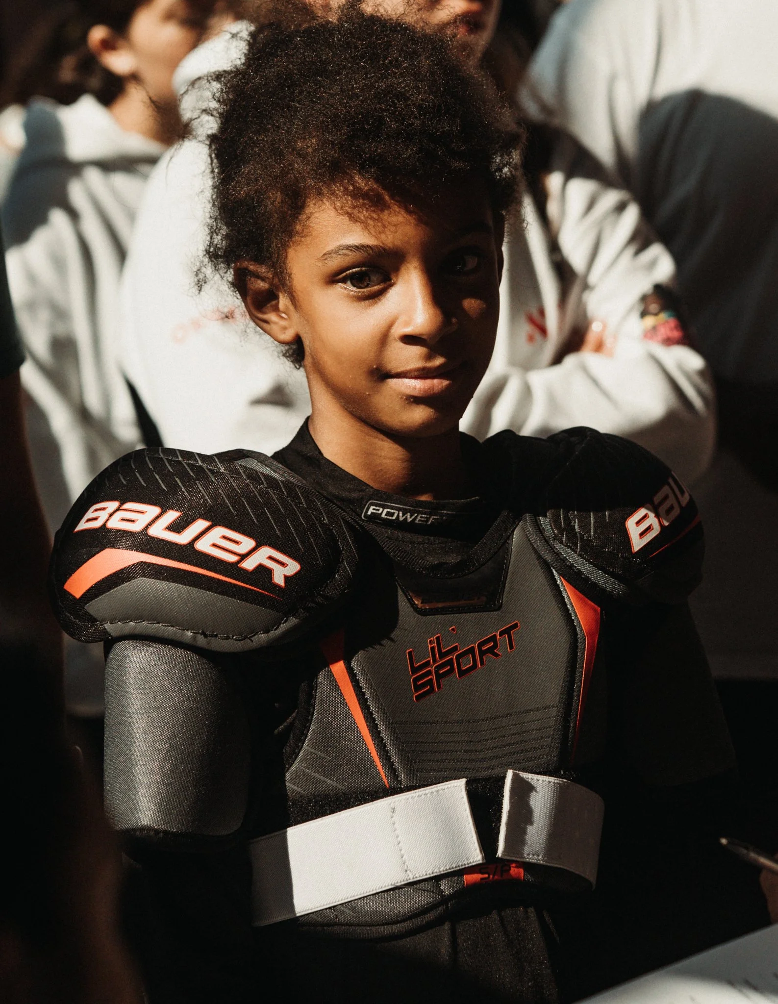 A young woman wearing black and red hockey gear, including shoulder pads, in a sports setting with people in the background.