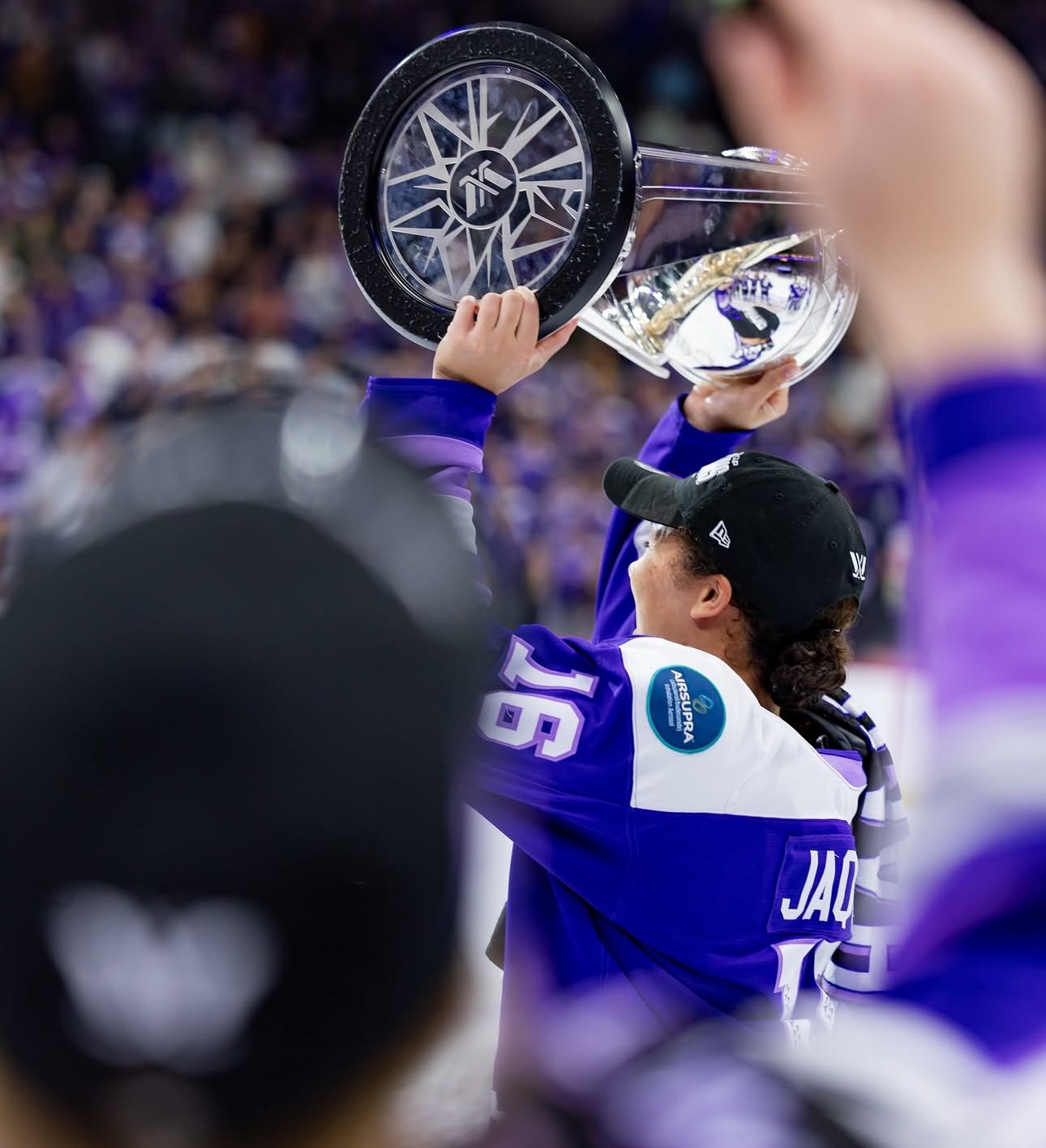 Hockey player wearing a blue jersey and black cap lifts a trophy above his head in celebration while surrounded by teammates and fans.