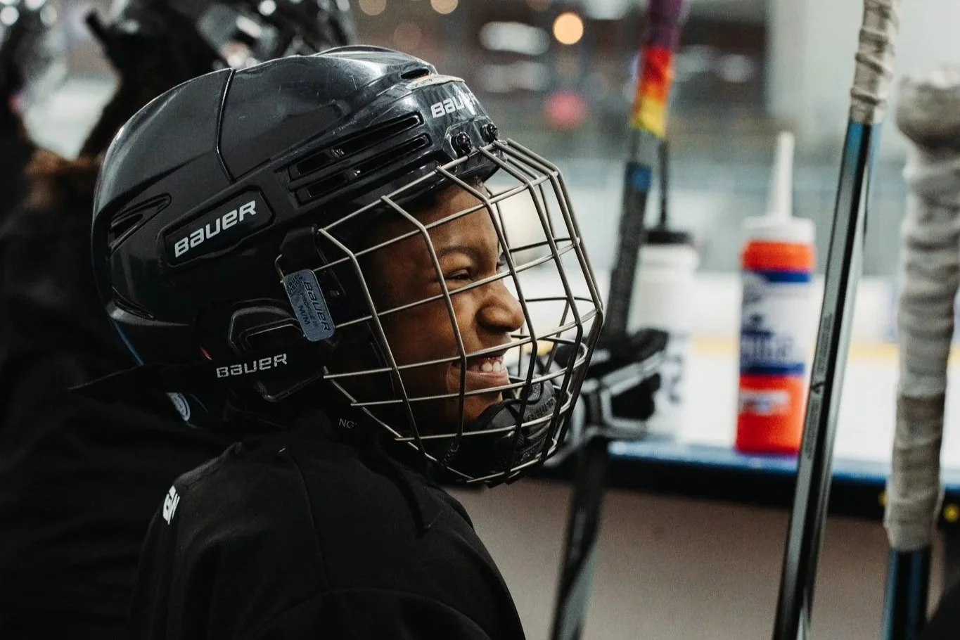 A female hockey player wearing a black Bauer helmet with a face cage, smiling between games on the bench