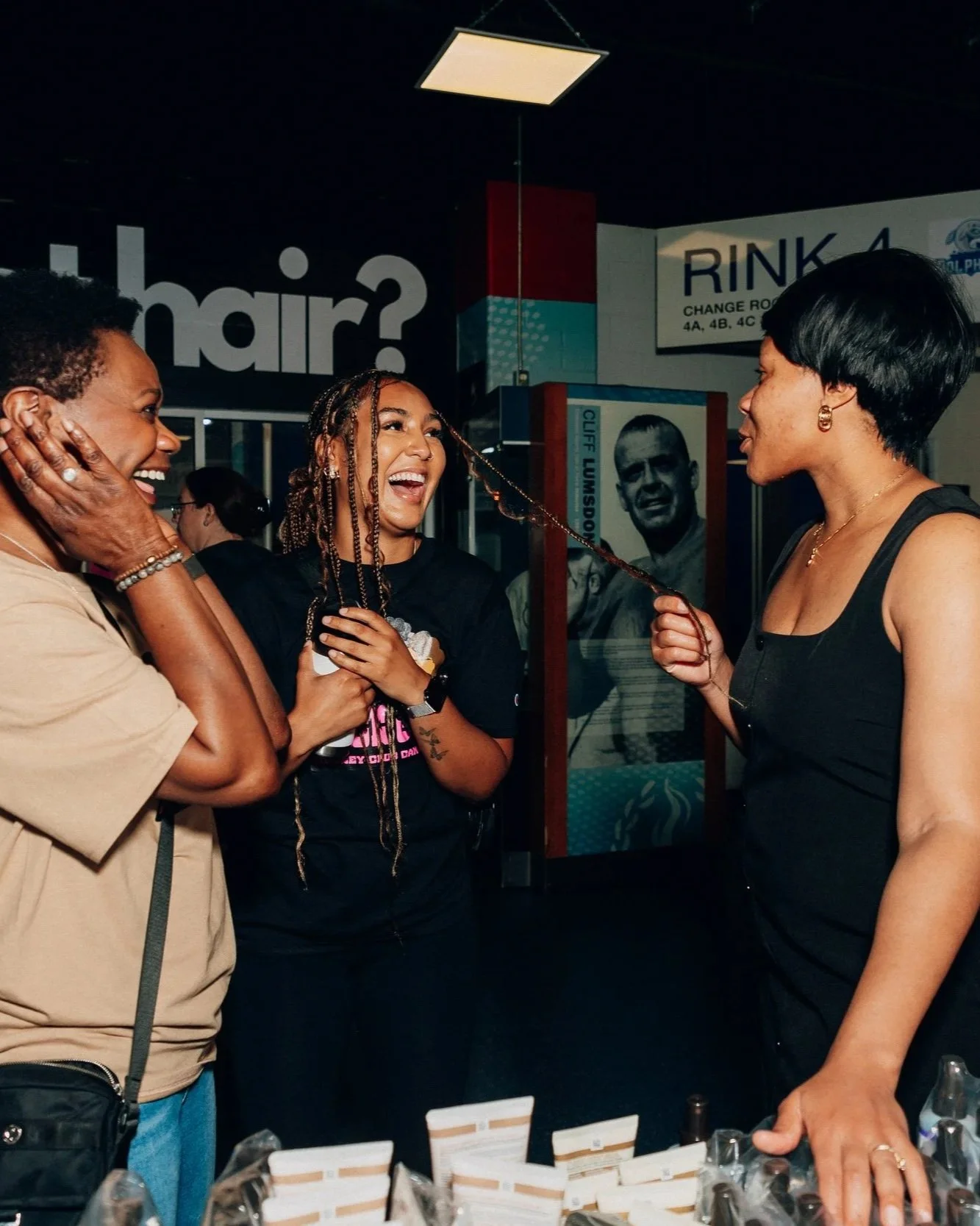 Three women laughing and talking at an indoor event, with a hair-themed background and a sign that says 'hair?'