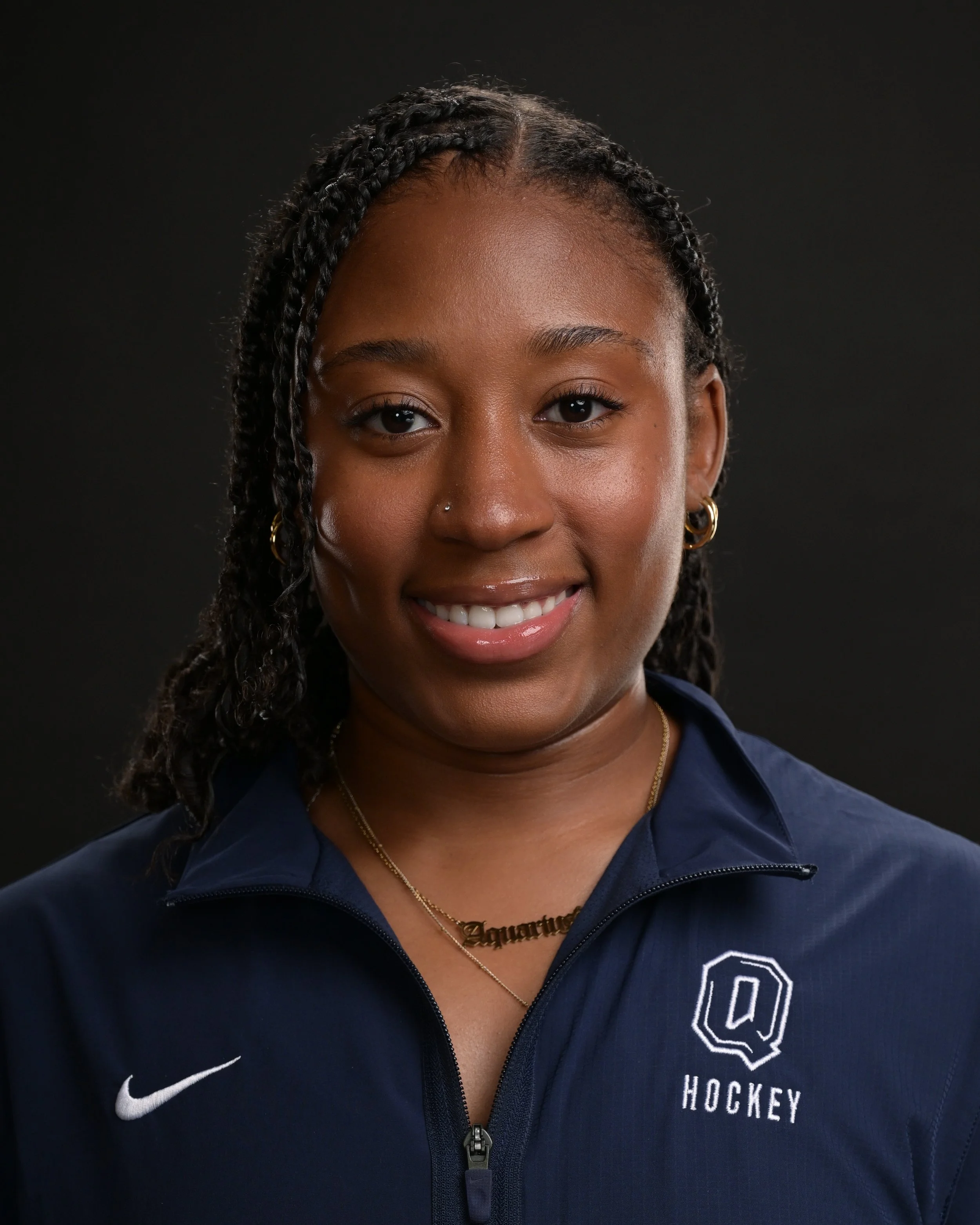 A young woman with braided hair, wearing a navy blue Nike jacket with a 'Q Hockey' logo, gold hoop earrings, a gold necklace, and a nose piercing, smiling against a black background.