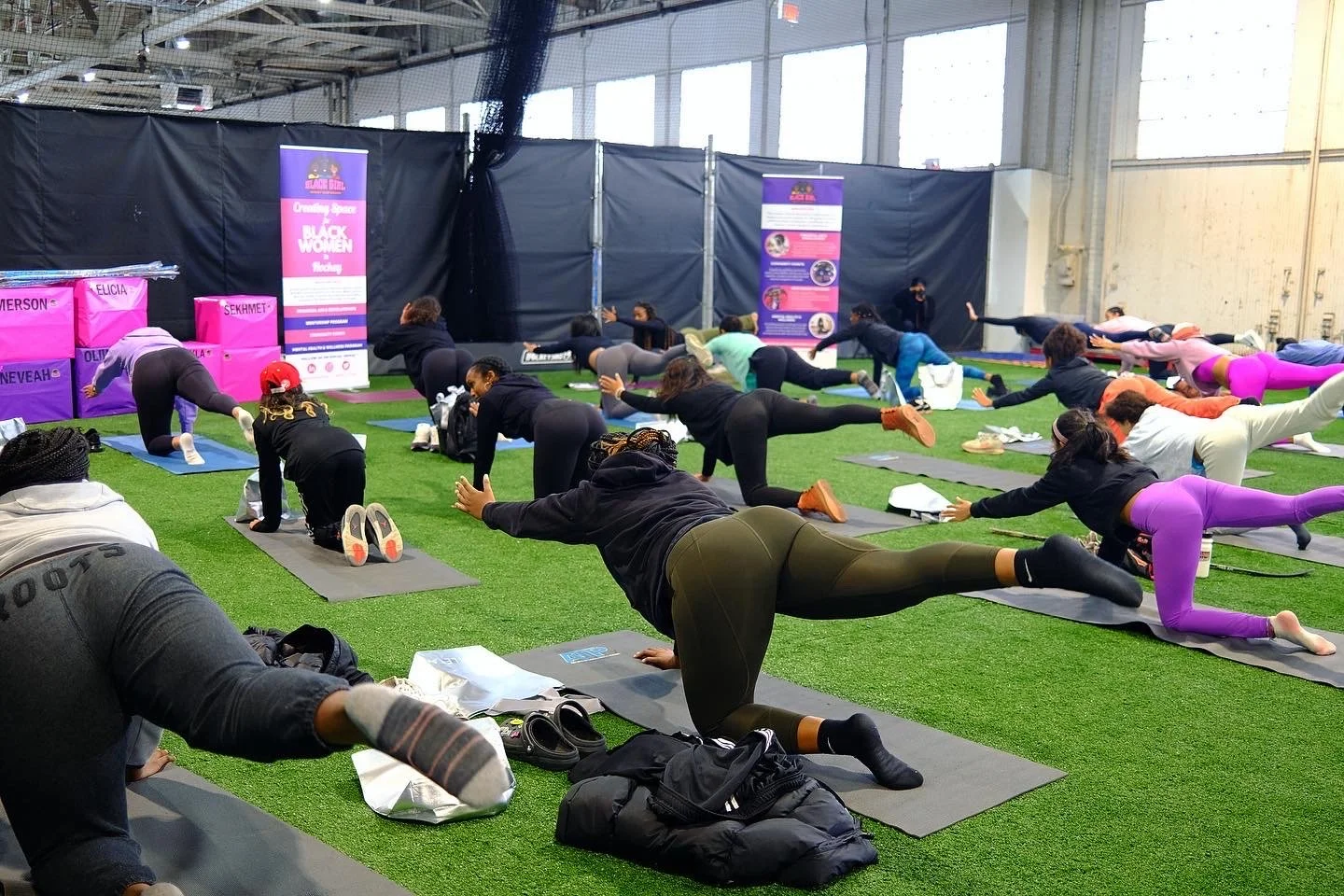 Group of people participating in a yoga or stretching class on gray mats, with some wearing workout clothes in purple and pink, inside a large indoor space with windows and black curtains.