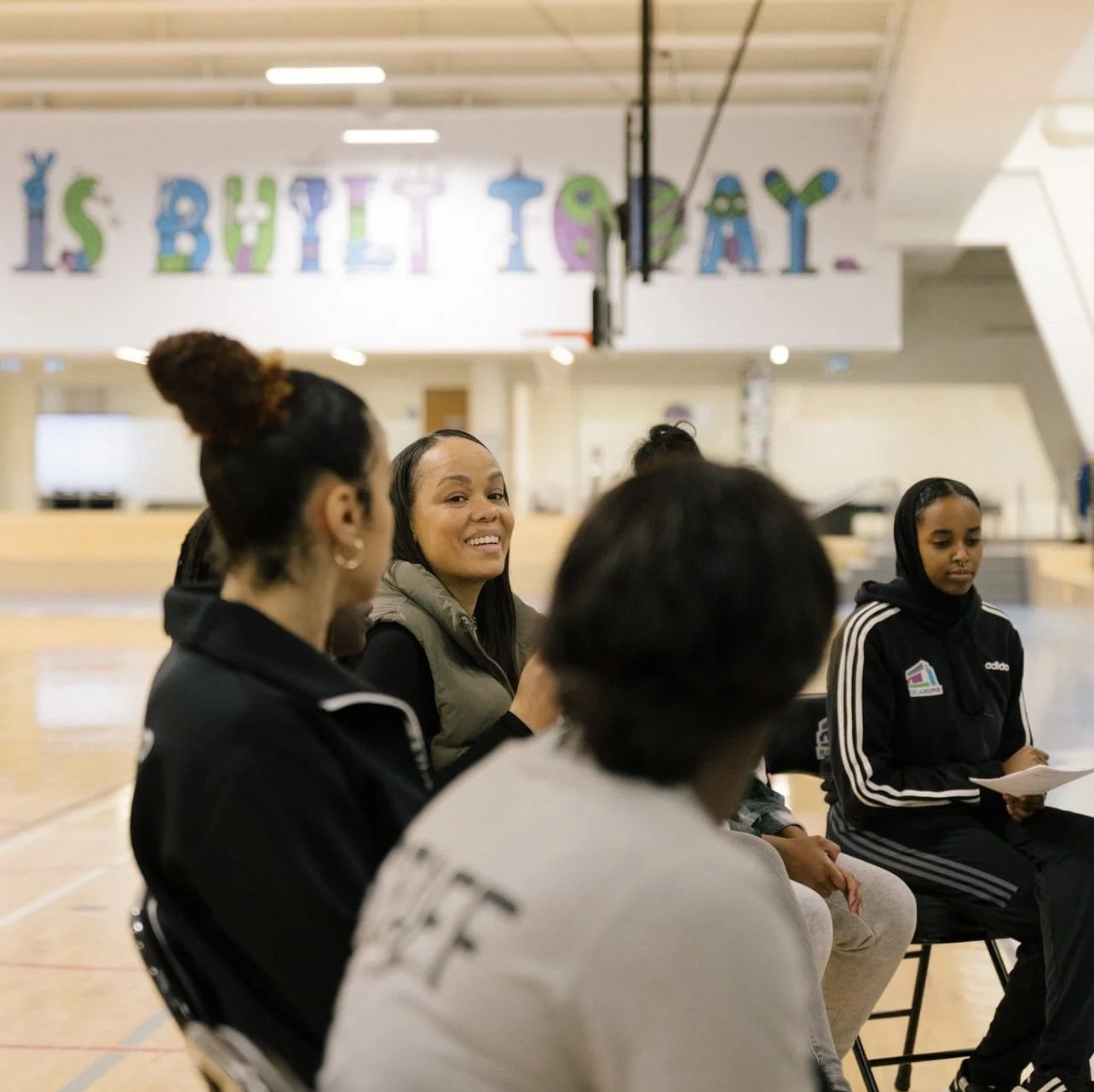 Group of women sitting and talking in a gymnasium with a birthday banner in the background.