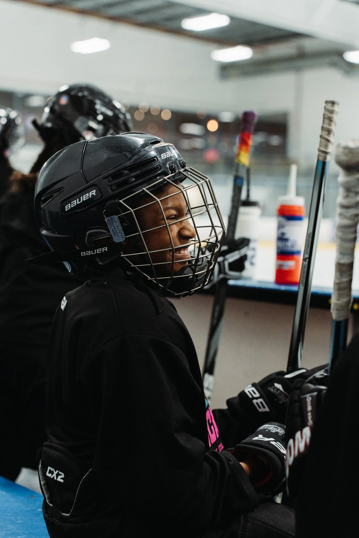 A young female ice hockey player on the bench smiling, wearing a black helmet and black jersey, with hockey sticks and supplies nearby in an ice rink.