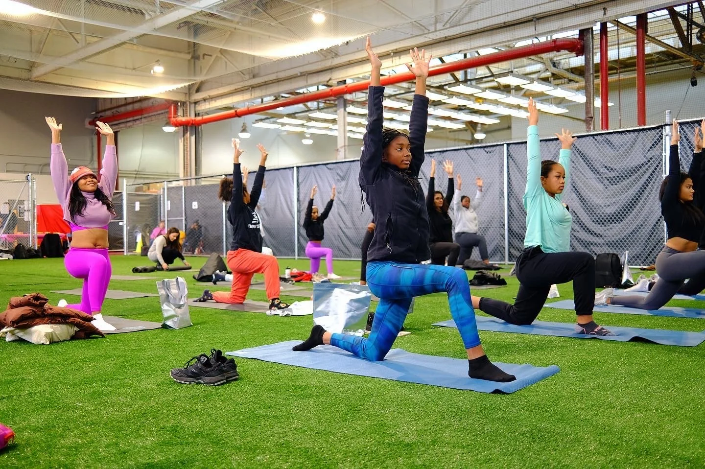Group of women doing yoga poses on mats indoors, with some kneeling and raising their arms overhead, in a spacious, well-lit facility with artificial turf flooring.